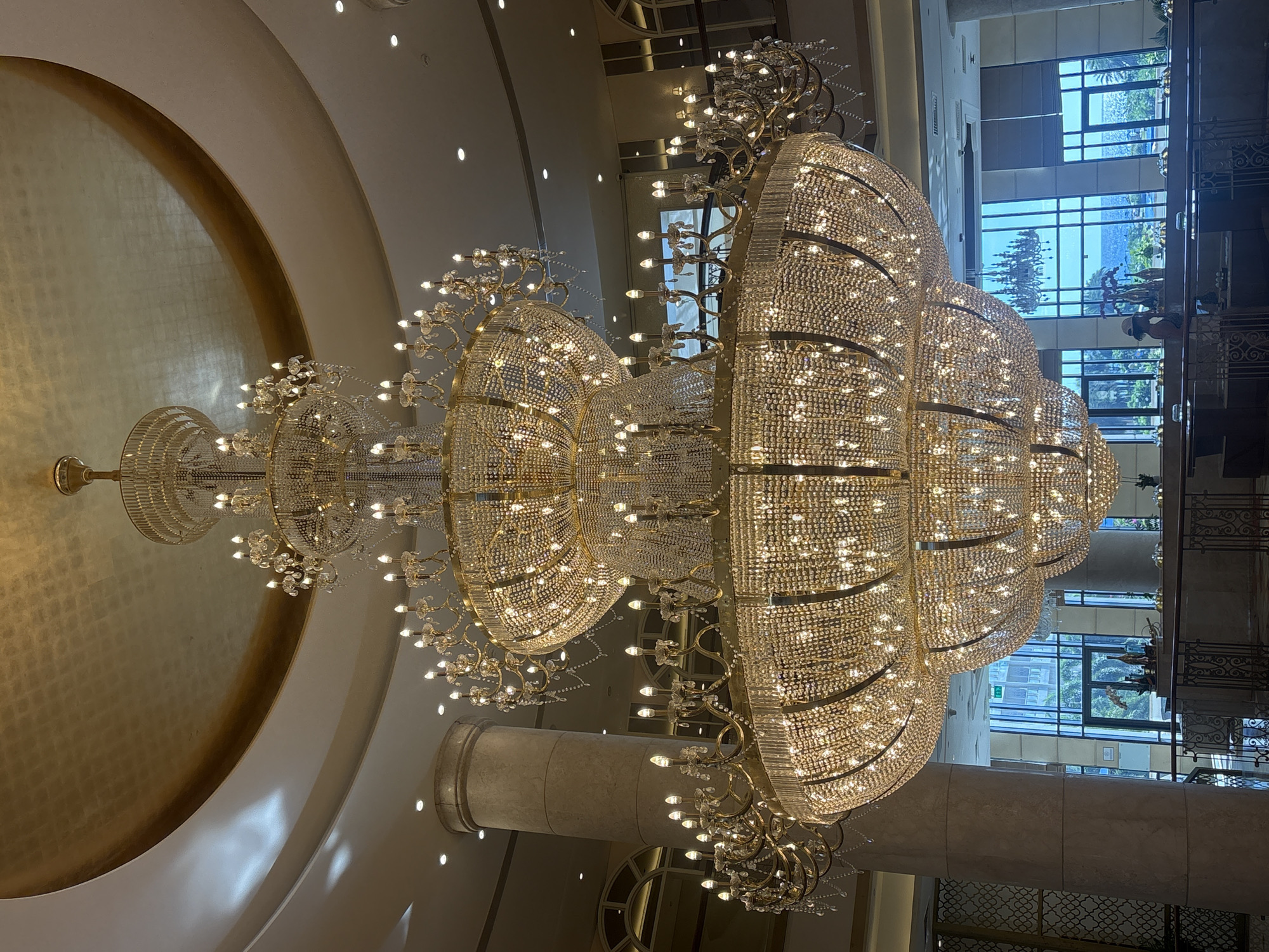 The image displays a grand, multi-tiered crystal chandelier hanging in what appears to be a hotel lobby or event space. The chandelier is illuminated and adorned with numerous crystals that reflect light. Above the chandelier, a curved ceiling is finished in a textured gold material. To the left, a large, fluted marble column is visible. In the background, through large windows, a view of the city of Đà Nẵng, Vietnam, is presented, featuring buildings and the ocean. A small number of people are partially visible in the background, near the windows, appearing to be guests or staff. The overall impression is one of opulence and spaciousness.