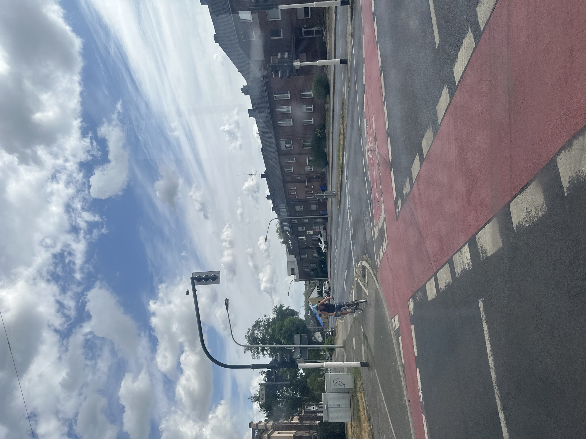 The image captures an outdoor scene on a cloudy day in Krefeld, Germany. In the foreground, a red-painted pedestrian crossing intersects with a road. A person on a bicycle, wearing a blue helmet and a dark tank top, is cycling across the intersection from left to right. In the background, a row of brick apartment buildings lines the street, with trees and other urban elements interspersed. Traffic lights and street lamps are visible, indicating a city environment. The sky is filled with white and grey clouds, suggesting dynamic weather conditions.