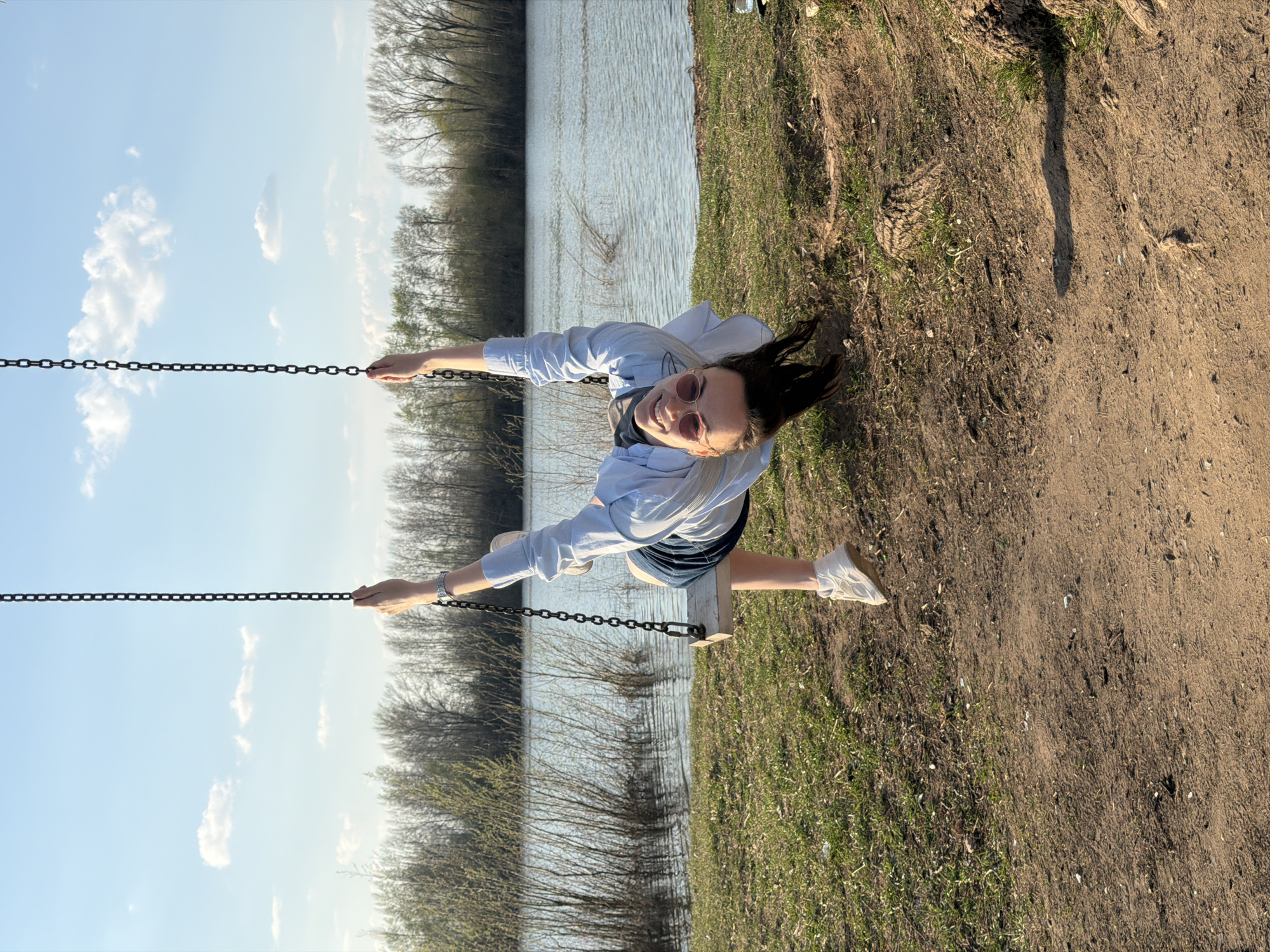 A person on a swing is centered in the image, against a backdrop with a body of water on one side and dirt/grass terrain on the other. The swing is attached to chains which the individual is holding. The water on the left side reflects the sky which is partially visible through the branches of surrounding trees. The dirt/grass terrain on the right side is visible and the sunlight is highlighting some of the ground. The individual is wearing sunglasses, a button up shirt, and sneakers. The location is likely near a body of water and a recreational area with the swing, possibly in Chaikovsky, Russia.