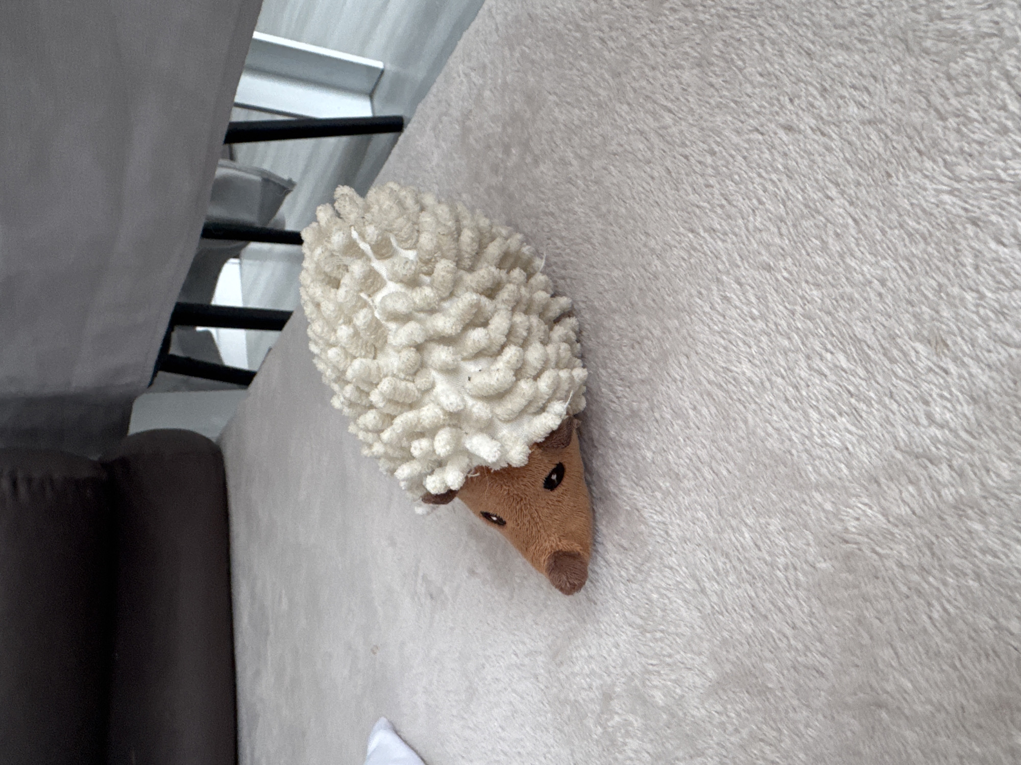 An indoor scene, located in Sofia, Bulgaria, features a plush toy hedgehog positioned on a light beige, shaggy rug. The hedgehog presents a brown, tapered snout with two small, black embroidered eyes and a nose. Its body is constructed from a dense, textured cream-colored fabric composed of numerous small, tightly clustered loops, simulating a spiny appearance. In the upper left quadrant of the background, a dark brown, possibly upholstered, object is partially visible. To the upper right, the lower edge of a grey fabric, likely a tablecloth, hangs above dark-colored furniture legs. Beneath these, a light-toned floor and a section of a white wall or door frame are discernible. No human subjects, actions, or interactions are present.