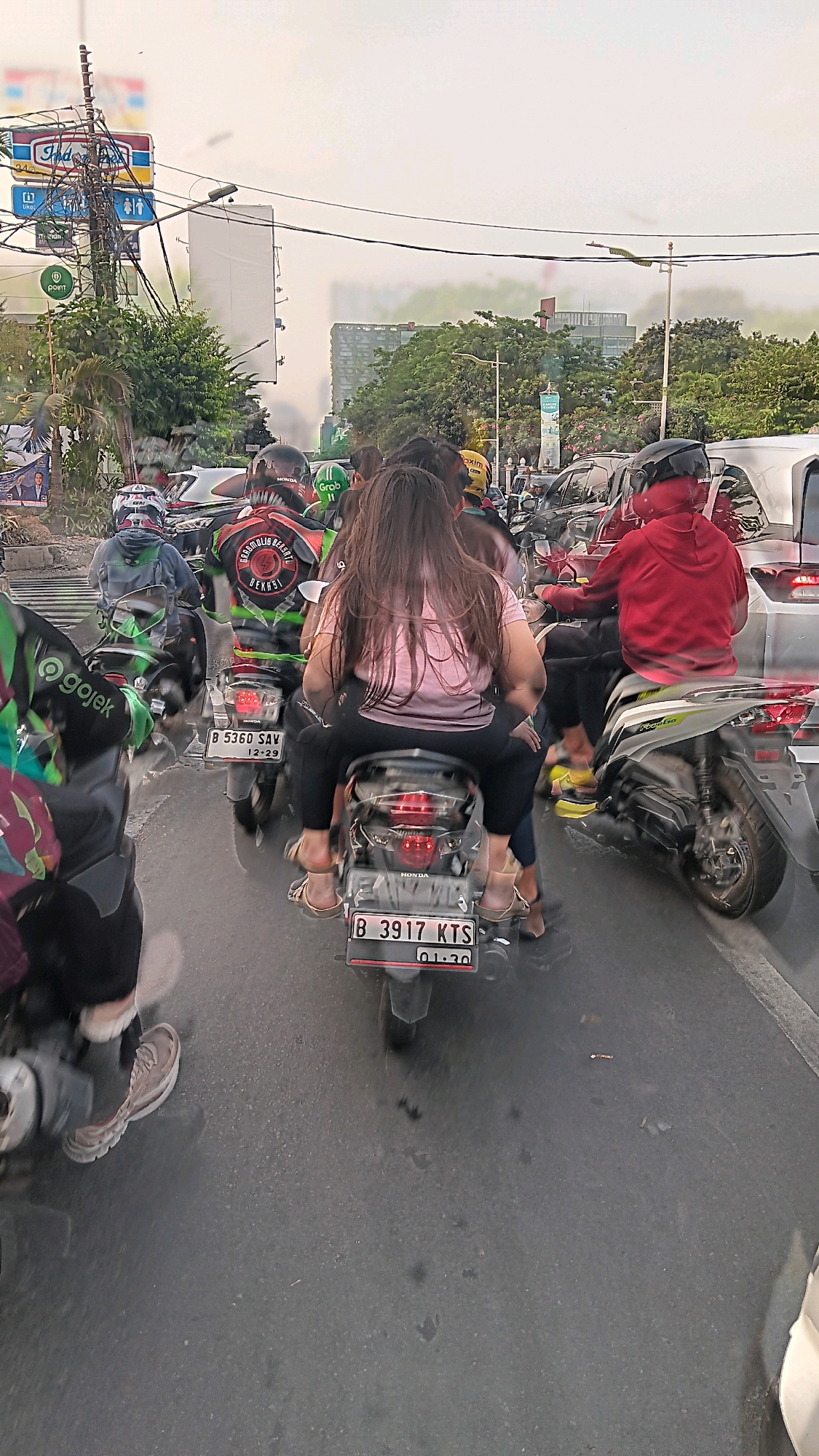 The image depicts a traffic jam in South Jakarta, Indonesia. Several motorcycles are visible, many carrying multiple passengers, a common sight in Indonesian urban traffic. The foreground shows the rear of a motorcycle with a license plate reading "B 3917 KTS". A woman with long brown hair is seated behind the driver, wearing a pink t-shirt and black pants. Her feet are visible, wearing sandals. To the left, a motorcycle with a "GoJek" logo is partially visible. In the middle ground, other motorcycles and cars are seen in congested traffic. The background shows trees, buildings, and overhead wires, indicating an urban environment. The sky is overcast, suggesting a cloudy day. The overall scene captures the dynamic and often chaotic nature of traffic in a bustling Indonesian city.