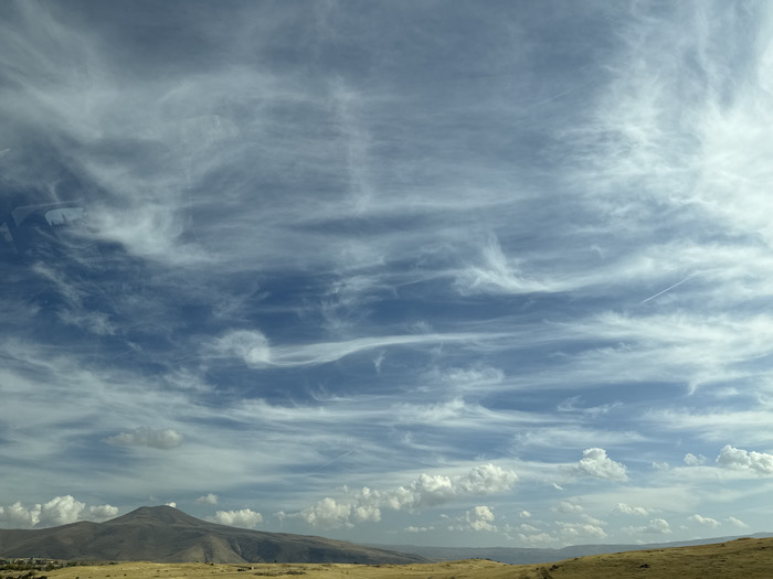 The image displays an expansive daytime landscape in Arzni, Armenia. The upper two-thirds of the frame are occupied by a deep blue sky, featuring numerous wispy white cirrus clouds stretching horizontally across the view and scattered cumulus clouds closer to the horizon. A faint white contrail from an aircraft is visible in the upper right quadrant of the sky. The lower third of the image shows an arid, undulating terrain covered in yellowish-brown dry vegetation. A prominent, dark-colored, conical mountain peak rises from the mid-ground on the left, with smaller, rolling hills extending towards the right horizon. No people or man-made structures are distinctly visible.