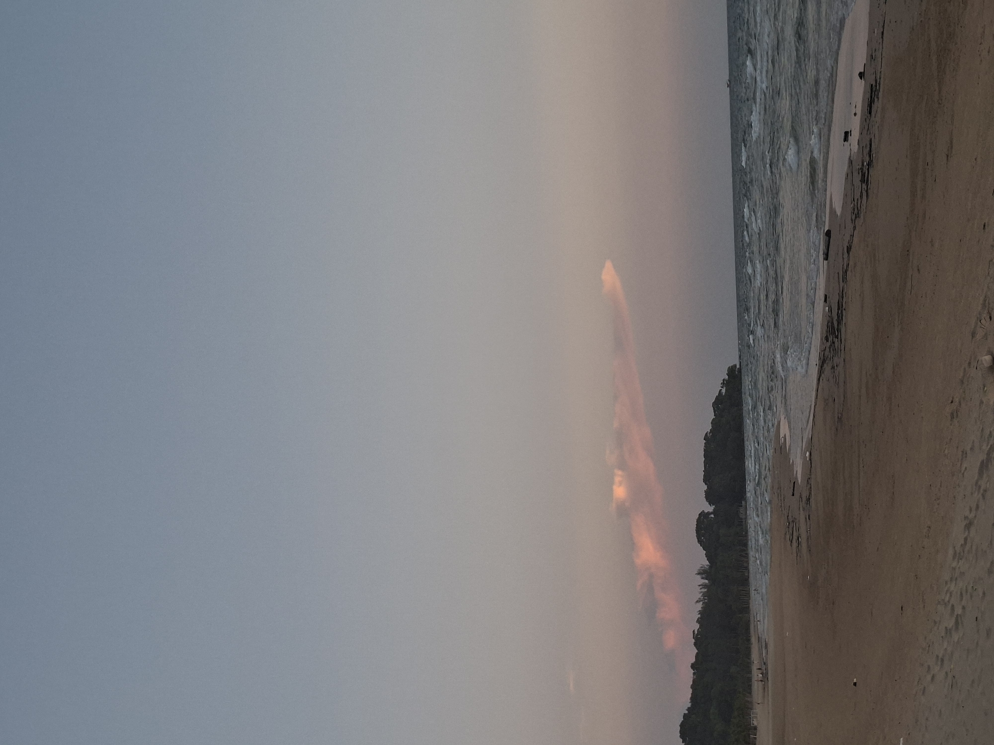 A serene, wide-angle shot captures a tranquil beach scene in Phú Quốc, Vietnam, during the soft light of dusk. The expansive sky transitions from a pale blue at the top to a gentle, warm hue near the horizon, tinged with subtle pinks from the setting sun. A wispy, elongated cloud, illuminated with a rosy glow, drifts across the sky. Below, a dense, dark green treeline forms the backdrop, suggesting a tropical landscape. The ocean stretches out, its surface rippled by moderate waves that break onto the sandy shore, creating white foam. The beach itself is a broad expanse of light brown sand, with some scattered debris, including pieces of driftwood and what appear to be small pieces of plastic. In the distance, on the left side of the beach, a few indistinct figures, likely people, are visible, suggesting a sparse presence of visitors. The overall atmosphere is one of peaceful isolation and natural beauty.