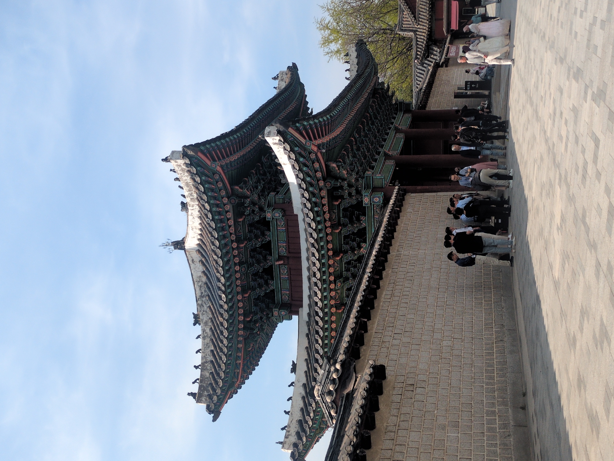 The image captures a low-angle shot of a traditional Korean building in Seoul, South Korea, under a bright sky. The building exhibits ornate, multi-tiered roofs with intricate carvings and colorful details. A stone wall forms the base of the structure, and dark wooden columns support the roofs. Several people are present in the scene, including a group dressed in traditional Korean clothing. People line up along a paved walkway near the building, while others stand in smaller groups. The environment suggests a historic or cultural site, possibly a palace or temple complex.