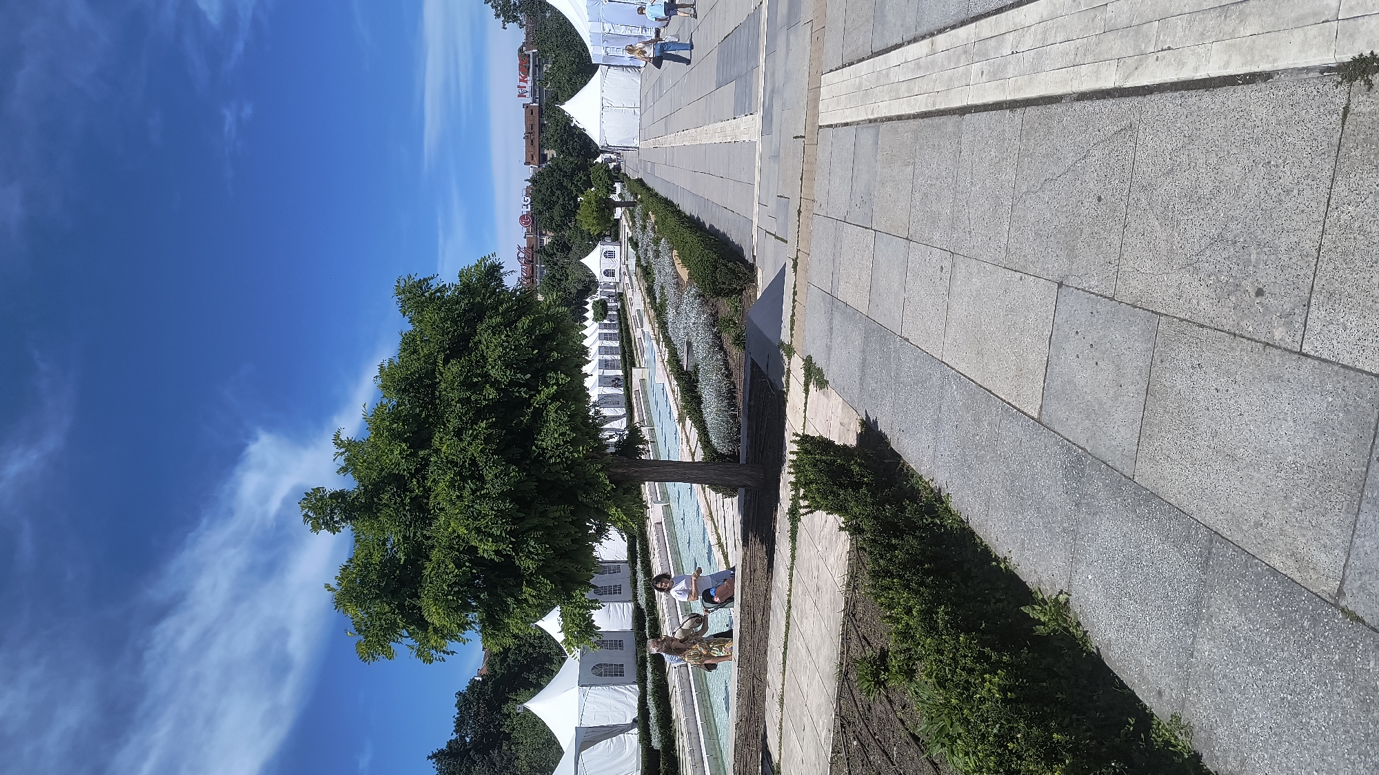 The image shows a long, stone-paved pathway with rows of identical white tents on the left side. A tree is visible on the left, casting a shadow onto the pathway. Two people are sitting on a stone bench near the tree. On the right side of the path are greenery and a paved area. The sky is blue with some clouds. The location is likely in a park or open space in Sofia, Bulgaria, possibly near a fair or event.