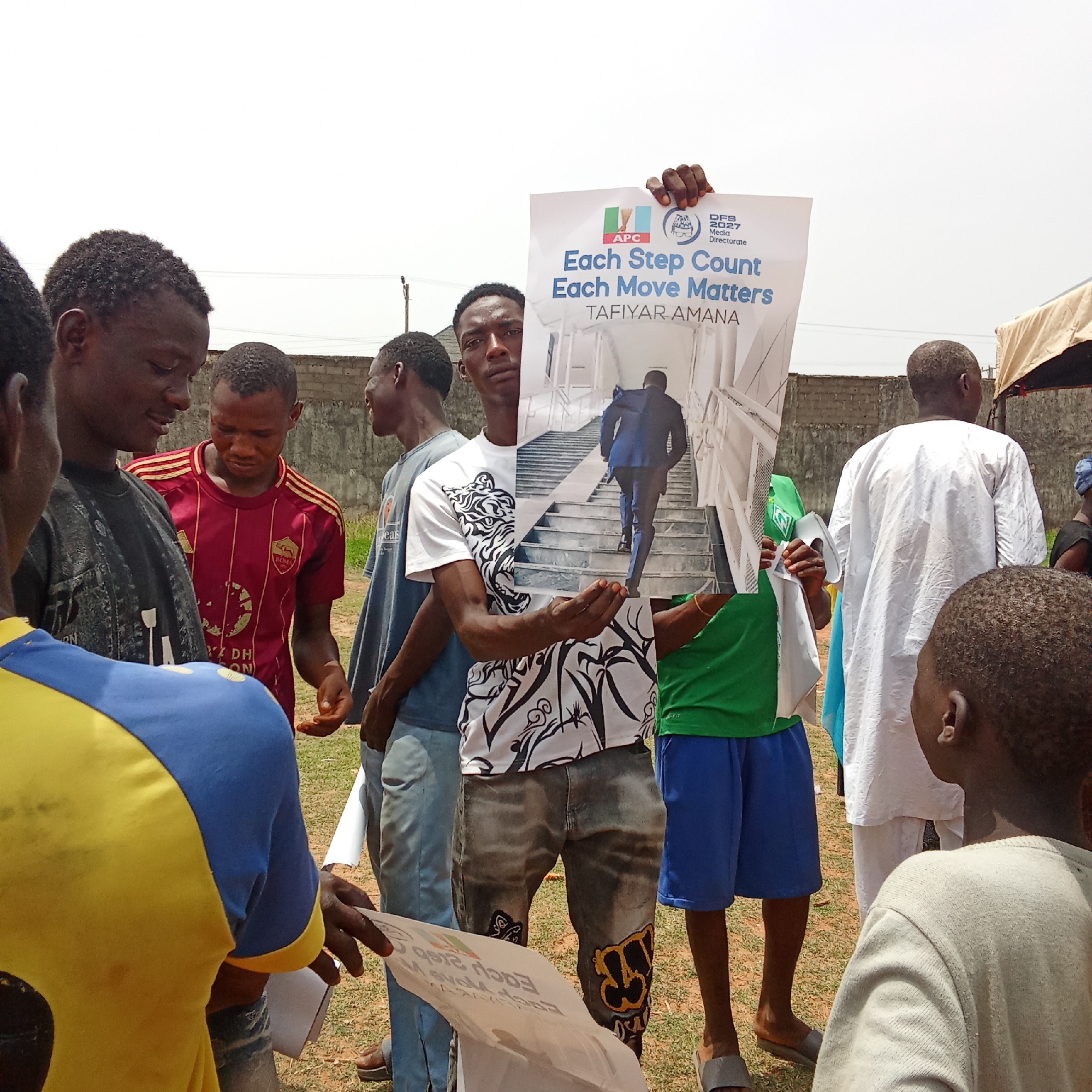 A group of approximately seven Nigerian men are gathered outdoors in Akwanga, Nigeria, under bright daylight. The central figure, a young man wearing a white t-shirt with a black and white design and distressed jeans, holds up a large political poster. The poster features a prominent image of a man in a blue suit ascending a staircase.
Visible text on the poster includes: "APC", "DFS 2027 Media Directorate", "Each Step Count Each Move Matters", and "TAFIYAR AMANA". The "APC" logo (All Progressives Congress) identifies the political party.
Around the poster-holder, other men are seen. To his left, another young man in a red and maroon "ROMA" football jersey looks down at something in his hands. Further left, a man in a dark t-shirt and another in a blue and yellow t-shirt are partially visible, also seemingly engaged with papers or discussion. To the right, a man in a green t-shirt and blue shorts is partially obscured by the poster, while another man in a traditional white garment (possibly an agbada) stands with his back to the viewer. A young boy's head is visible in the foreground on the far right.
The setting appears to be an open area, possibly a field or a compound, with a low concrete block wall and some sparse vegetation in the background. A light-colored tent or canopy is partially visible on the far right. The clear, bright sky suggests sunny weather. The gathering appears to be a casual political outreach or campaign event, with individuals distributing or discussing campaign materials.