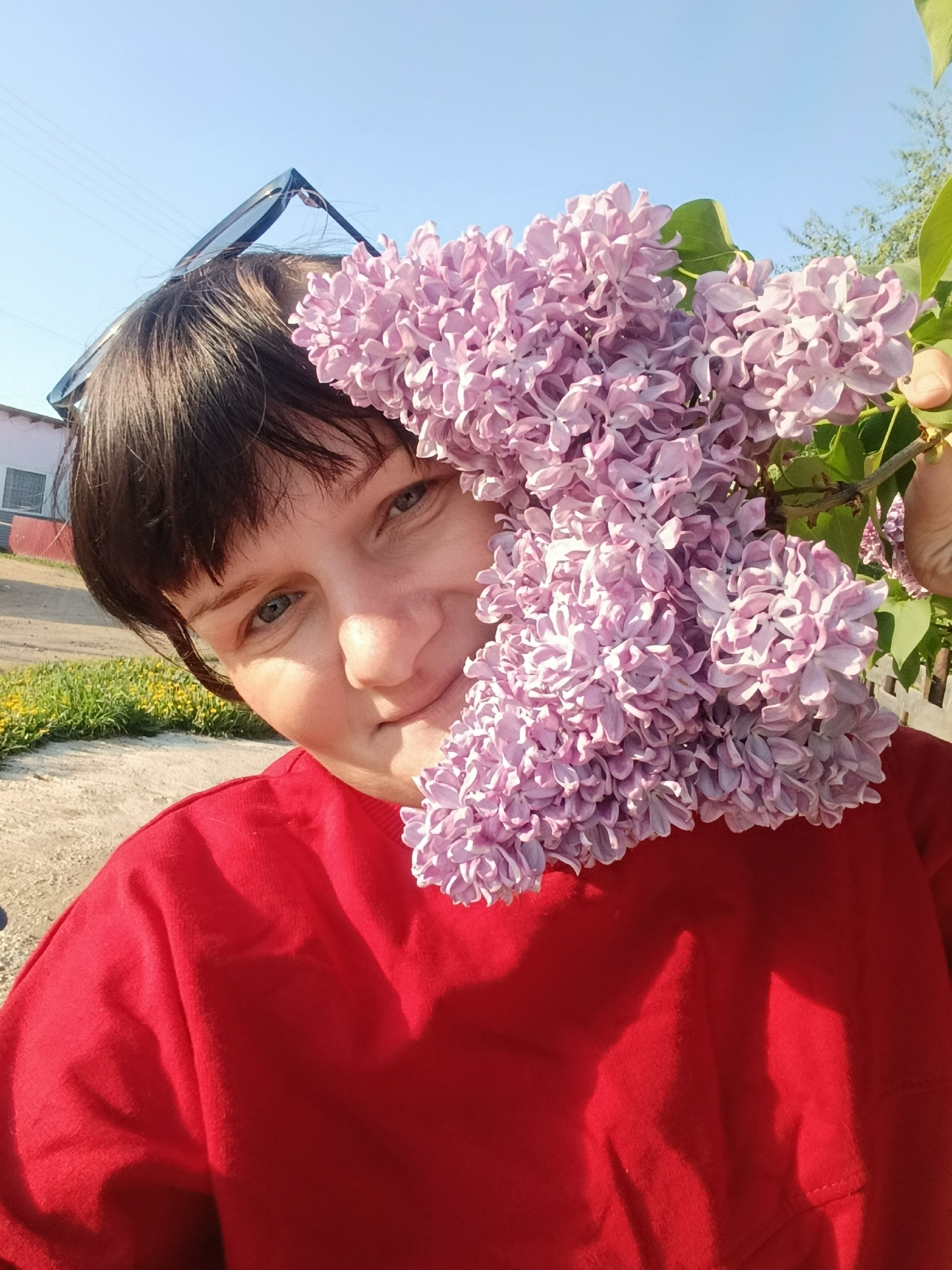 A woman with short dark hair and blue eyes is holding a large bunch of lilac flowers in front of her face. She is wearing a red long-sleeved shirt. The flowers are a light purple color. The background includes a gravel road, grass, a building with a blue roof, and a white fence. The picture appears to be taken outdoors in daylight, possibly in the village of Фоки, Russia based on the metadata.