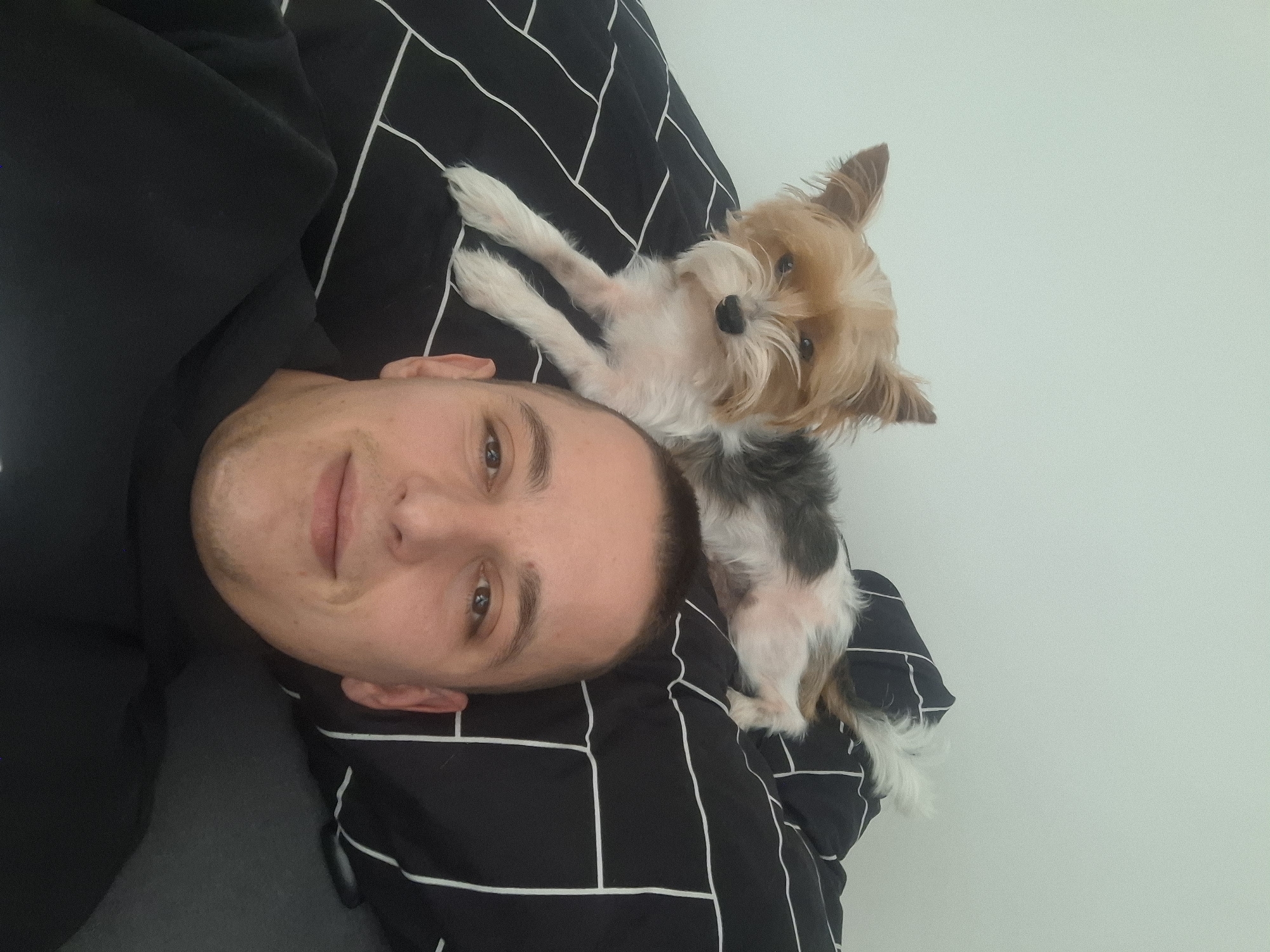 The image depicts a male individual and a small dog resting together in an indoor setting, specifically a bed, located in the city of Gunzenhausen, Germany. The man, positioned in the lower half of the frame, has short dark hair, light skin, and a subtle smile directed towards the camera. He is wearing a black long-sleeved garment, possibly a hoodie. Behind his head, on a pillow, a Biewer Yorkshire Terrier or similar tricolor dog (tan, white, and dark grey/black fur) is lying down, facing the camera with its front paws extended. The dog's ears are erect. The bedding consists of a black and white geometric patterned duvet cover or pillowcase featuring intersecting white lines on a black background. A plain, light-colored wall serves as the background above the bedding. A dark grey fabric is visible in the lower right corner. Both subjects appear to be relaxed.