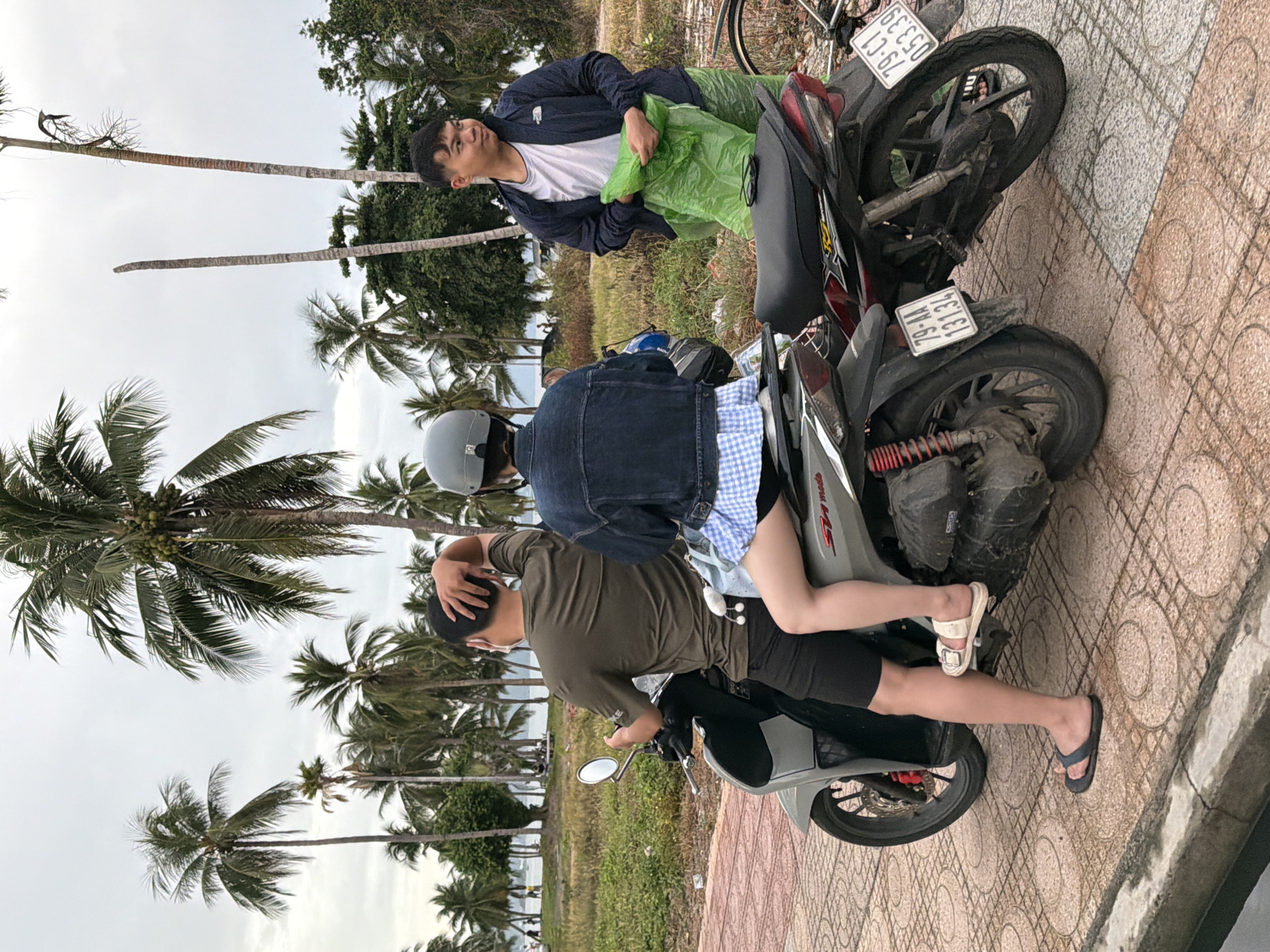 A scene captured on a cloudy day in Nha Trang, Vietnam, features three individuals and two motorcycles on a paved walkway lined with palm trees. A man, facing away from the camera and wearing a green t-shirt and black shorts, is seated on the left motorcycle. A woman, with a blue and white checkered skirt and a denim jacket, is seated behind him. To the right, another man stands holding a green plastic bag and a jacket. The motorcycles are parked side-by-side, with license plates visible. The background reveals a grassy field with more palm trees and a glimpse of the ocean in the distance, suggesting a coastal location.