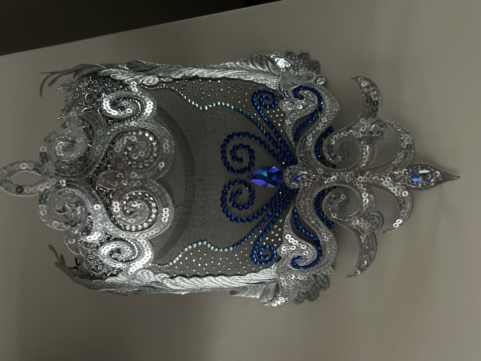 The image shows an ornate, rectangular fabric embellishment with a gray net backing. Silver sequins and embroidery form a detailed border and swirling patterns. Blue beadwork adds contrast around a large, multifaceted blue jewel in the center. The embellishment is mounted on a light-colored wall. There is no information in the image that connects it to the city of Moscow, Russia.
