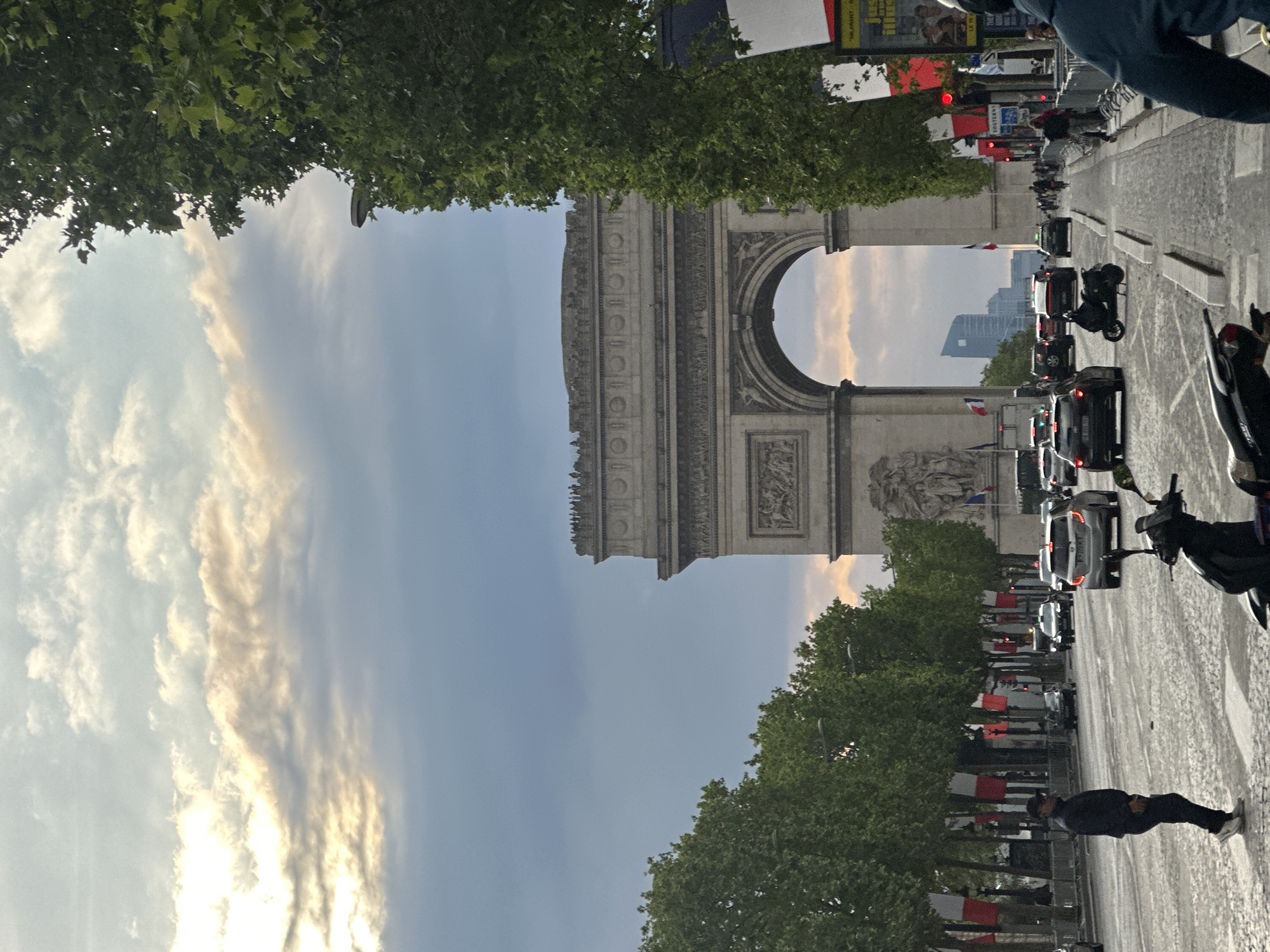 The image shows the Arc de Triomphe in Paris, France. Cars and motorcycles are lined up on the avenue leading towards the arch. There is a person walking on the sidewalk. French flags line the street. Trees are visible along the sides. The sky is cloudy. Buildings are visible through the arch.