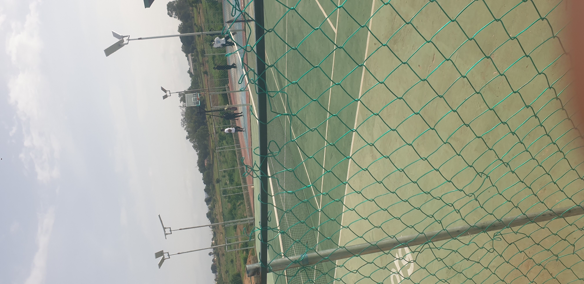 The image depicts an outdoor sports facility, likely in Jos, Nigeria, featuring a tennis court in the foreground, partially obscured by a green chain-link fence. Beyond the tennis court, a basketball court is visible with several individuals present. There are two men in casual attire standing on the basketball court, one in a white shirt and the other in a dark shirt. Another group of three individuals, also casually dressed, are near the basketball hoop. The surrounding environment includes lush green vegetation, trees, and a hilly landscape in the background under a hazy sky. Several tall lampposts with solar panels are strategically placed around the facility, suggesting it is equipped for evening use. The overall scene suggests a recreational area within a city or town.