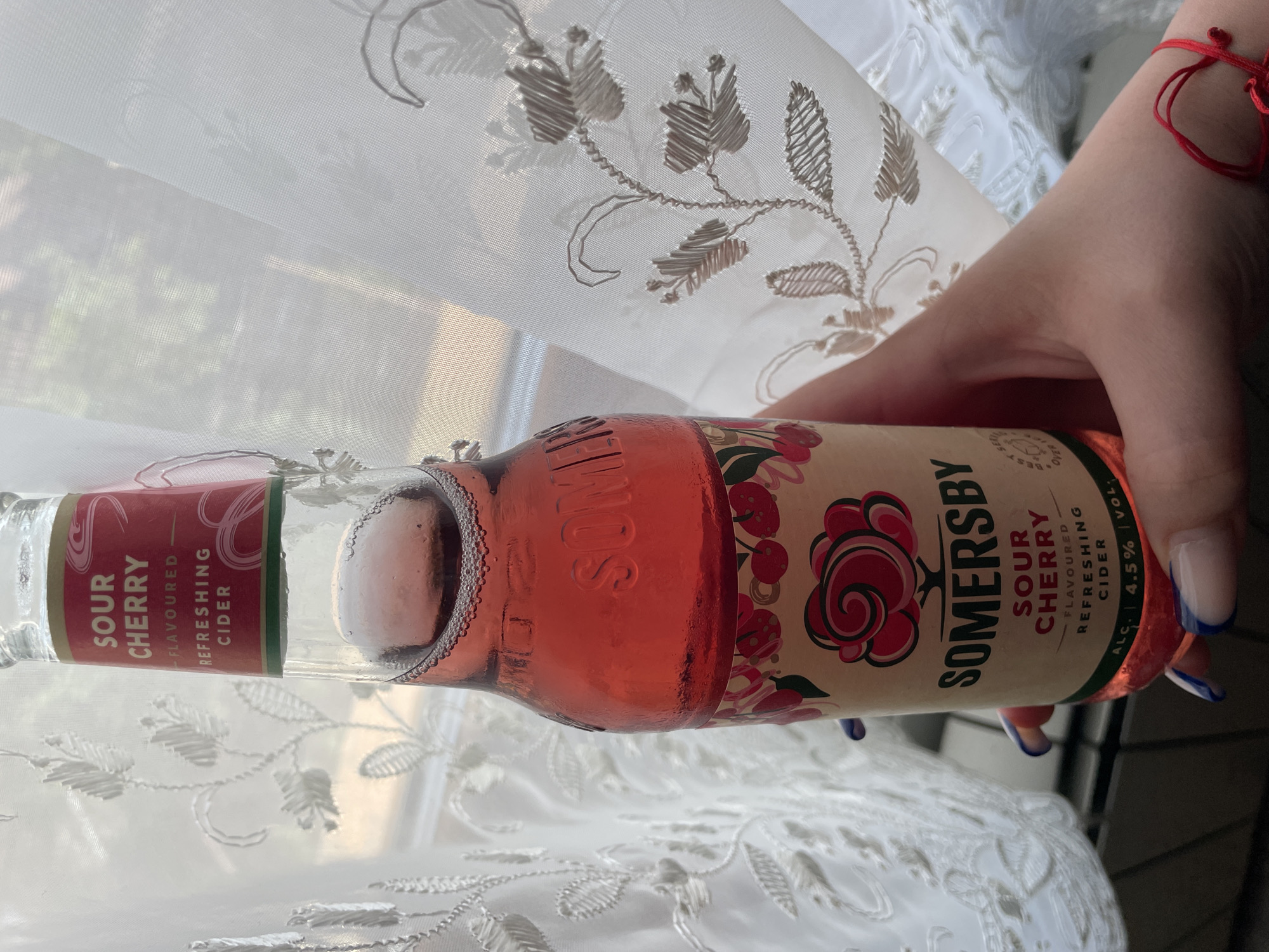A hand with blue-tipped nails is holding a bottle of Somersby Sour Cherry Flavoured Refreshing Cider. The bottle is partially obscured by a white curtain with a floral design. The background is softly illuminated. The location is not specified in the image.
