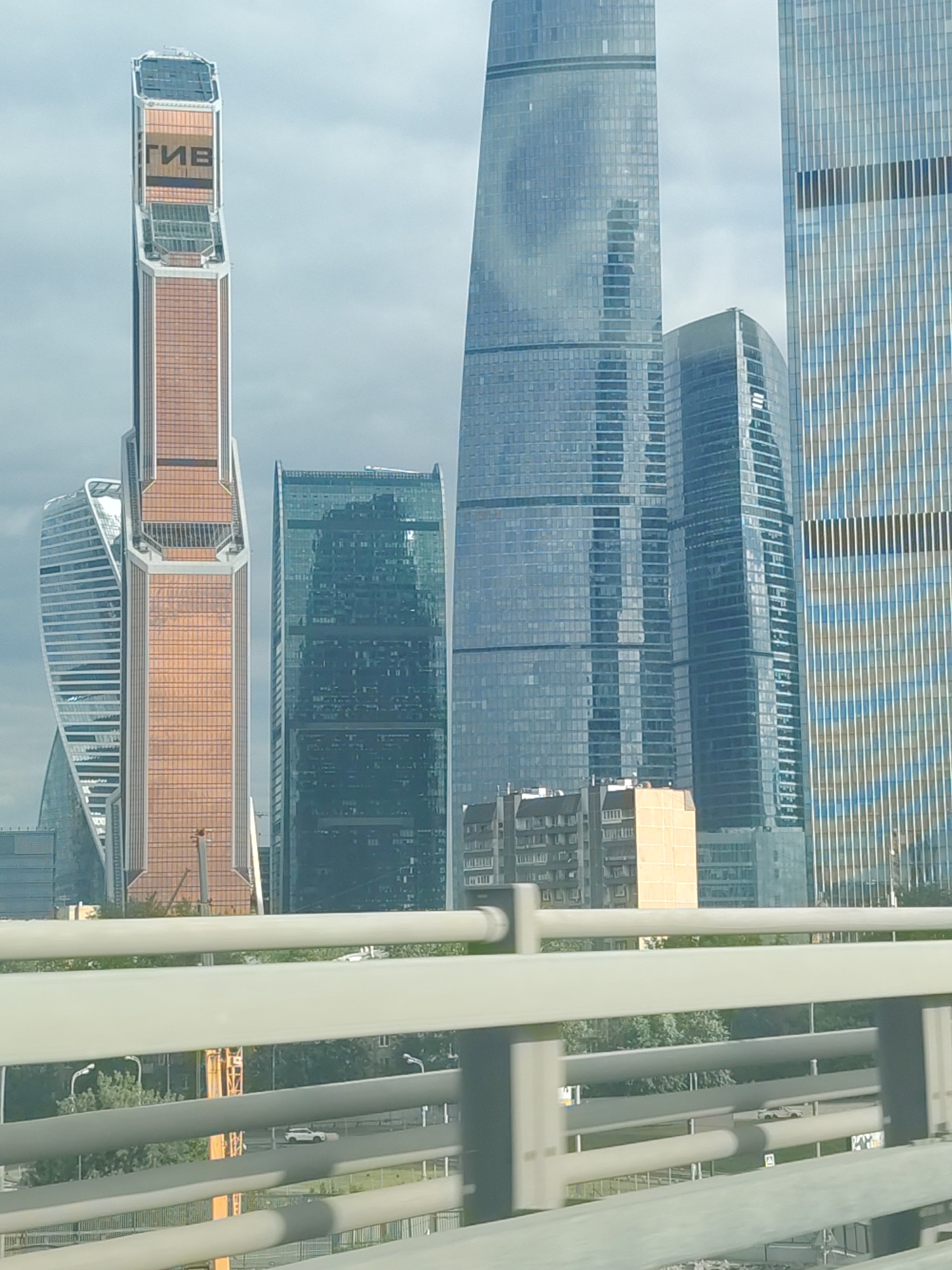 The image shows the Moscow International Business Center, also known as Moscow City, a business district under development in central Moscow, Russia. Several skyscrapers are visible, including the Neva Towers, Mercury City Tower, and Eurasian Tower. The weather is overcast. In the foreground, a white railing is prominent, obscuring a view of cars and trees on a lower level.