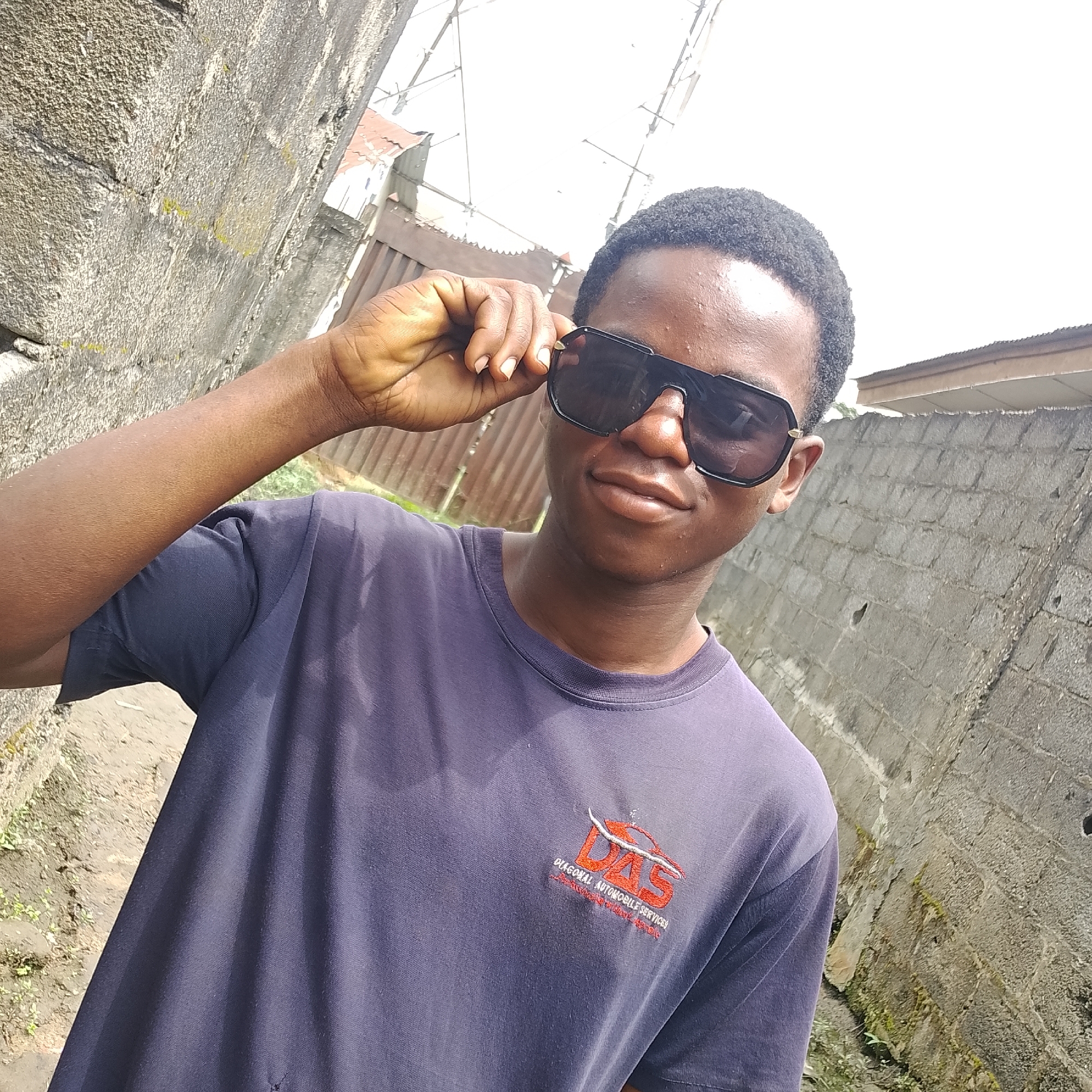 A young Black man is pictured from the chest up, facing slightly to the right, wearing dark sunglasses and a dark purple t-shirt. He is adjusting his sunglasses with his left hand. The t-shirt has an embroidered logo on the left chest that appears to be red and white, with text below it. He has short, curly black hair. The background consists of a rough, textured concrete wall on the right and a similar wall on the left, with a narrow dirt path between them. The sky above is bright white, suggesting daylight. Various metal structures and building roofs are visible in the distance between the walls.