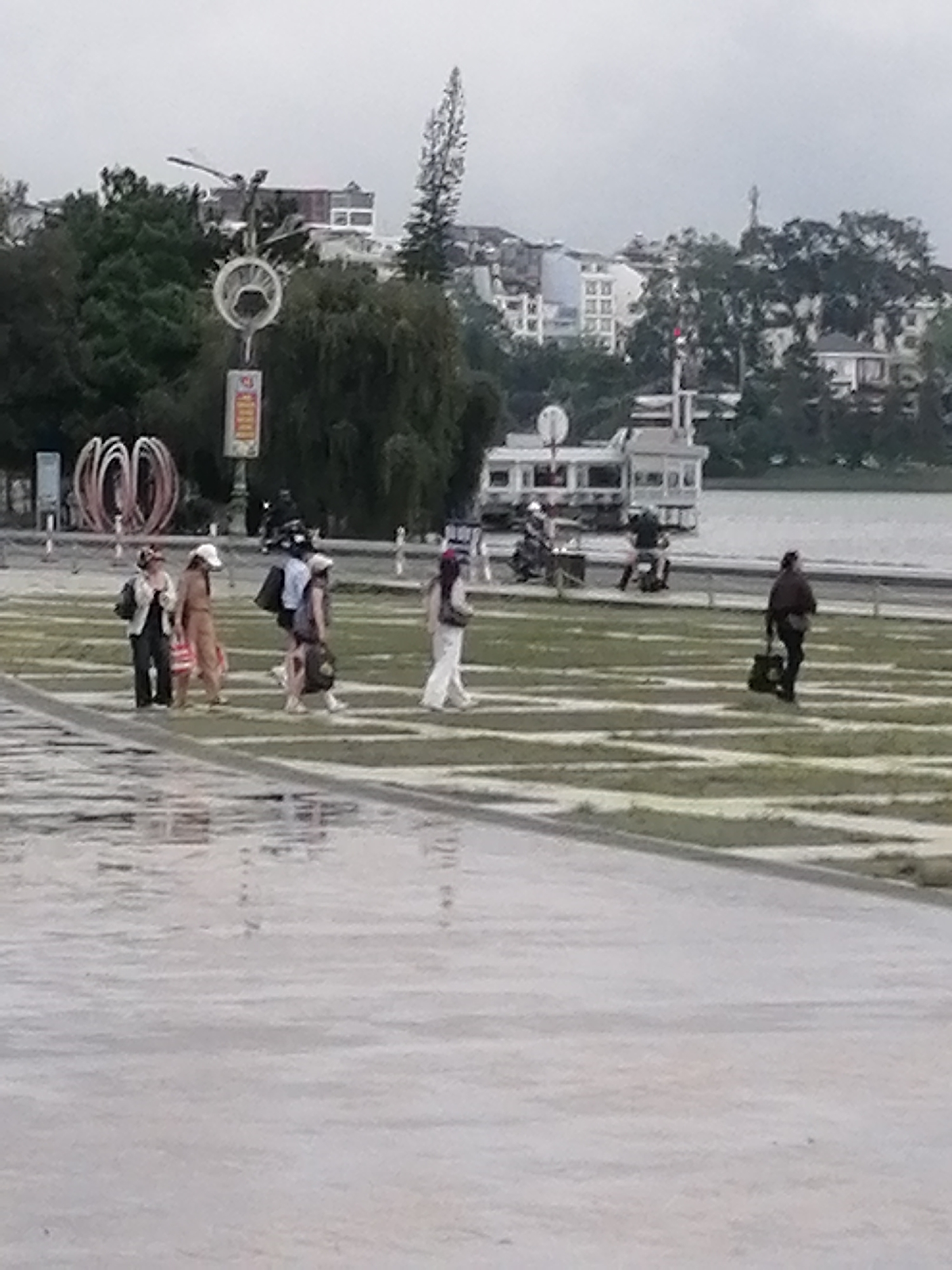 The image depicts a cloudy day at Xuan Huong Lake in Da Lat, Vietnam. In the foreground, a wet paved area reflects the overcast sky. Beyond this, a section of manicured grass with diagonal lines suggests a park or recreational area. Several people are walking across this grassy area.
On the left side of the grassy area, a group of four people are walking together. The person on the far left wears dark pants and a light jacket. Next to them, a person in a tan outfit walks with a bag. A person in dark clothing and a light hat follows, carrying a bag. The fourth person in this group wears a dark top and a lighter skirt, also carrying a bag.
Further to the right, a single person in white pants and a light top walks across the grassy area, carrying a dark bag over their shoulder. To the far right, another individual in dark clothing walks away from the camera, carrying a dark bag.
In the background, a white boat with a multi-level structure is docked on the lake. Beyond the lake, buildings and lush green trees form the city skyline. A tall, slender tree with a unique shape stands prominently amongst the other foliage. Several other indistinct figures and objects are visible in the midground, including what appear to be motorcycles. A circular decorative structure with multiple rings is visible in the left background, near a signpost. The overall impression is of a public space being utilized by pedestrians on a subdued day.