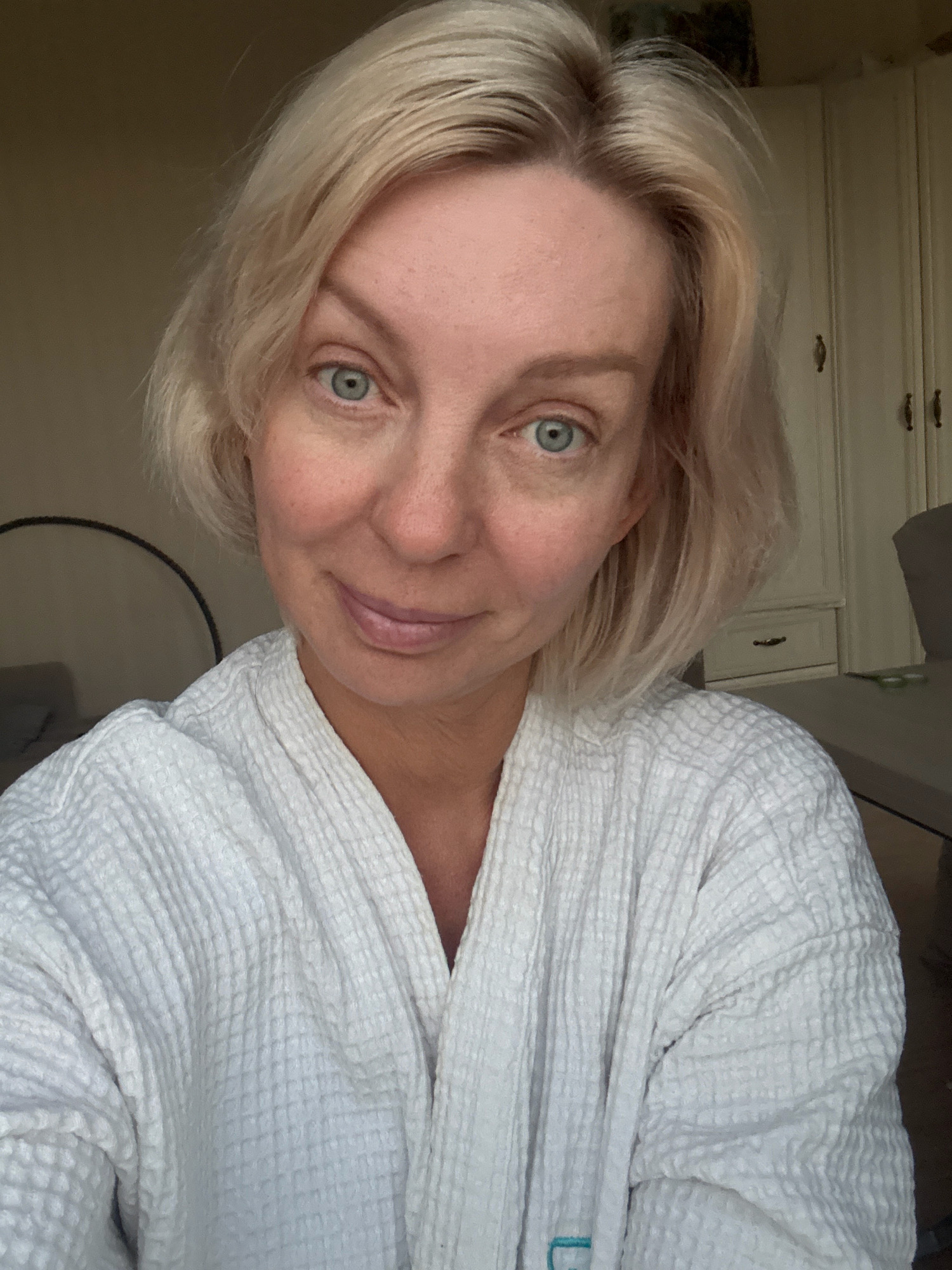 A close-up, selfie-style photograph shows a woman with short, blonde hair and blue eyes. She is wearing a white waffle-weave robe. The background appears to be an indoor setting, possibly a bedroom, with a wardrobe and a table visible. The lighting suggests it may be daytime. The image does not contain any contextual information that would allow for determining a specific location in Sochi, Russia.