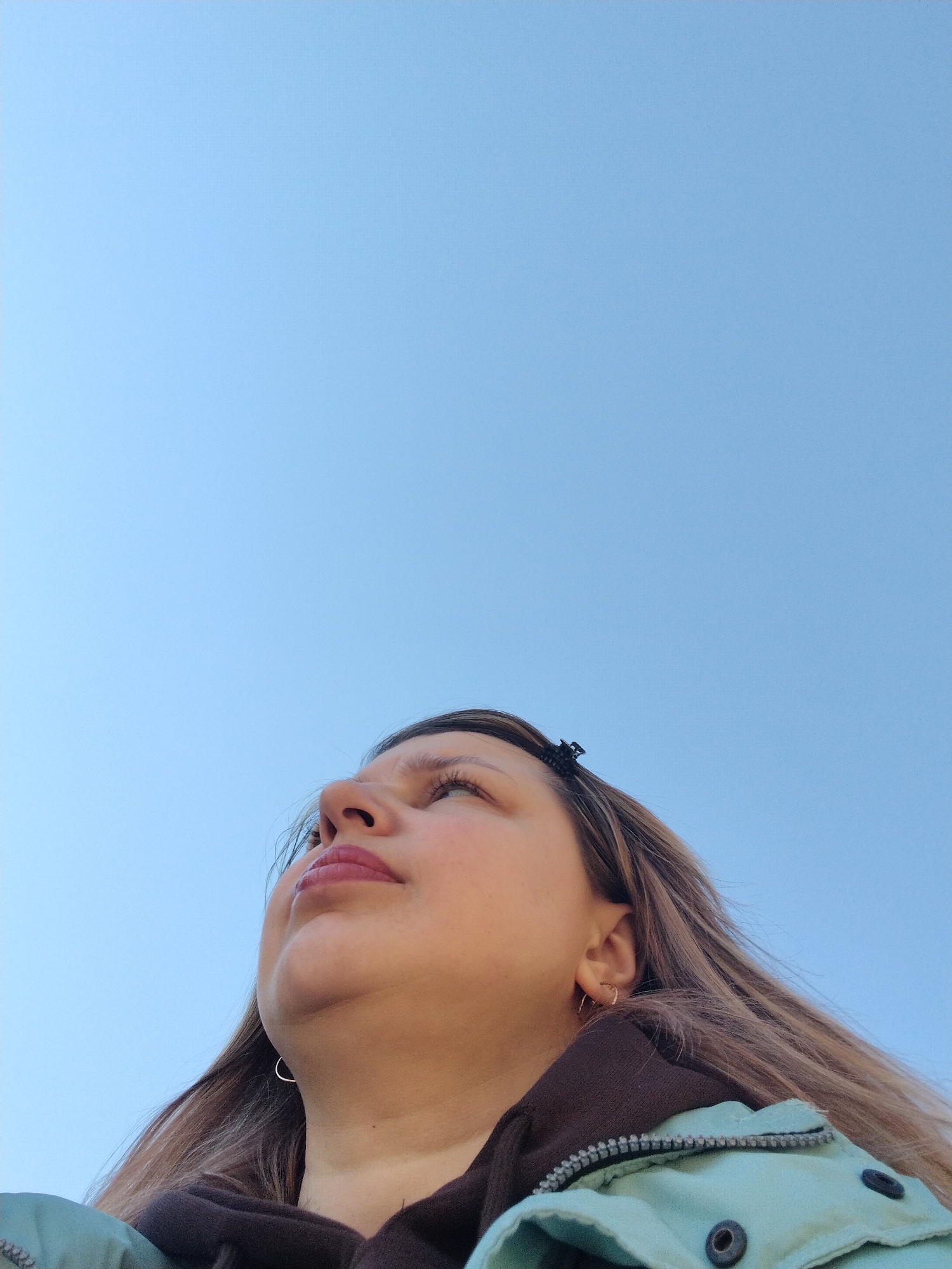 A low-angle shot captures a woman against a clear, pale blue sky. The woman, positioned in the lower portion of the frame, is looking upwards. Her face is illuminated, revealing her features: a slightly upturned nose, full lips, and light-colored eyes. She has medium-length brown hair. A small, dark hair clip is visible near the top of her head. She is wearing a light blue jacket over a brown hoodie. Silver hoop earrings are visible in her ears. The location is Rusanovo, Russia. There is no indication of location within the city, this scene could be anywhere within Rusanovo.