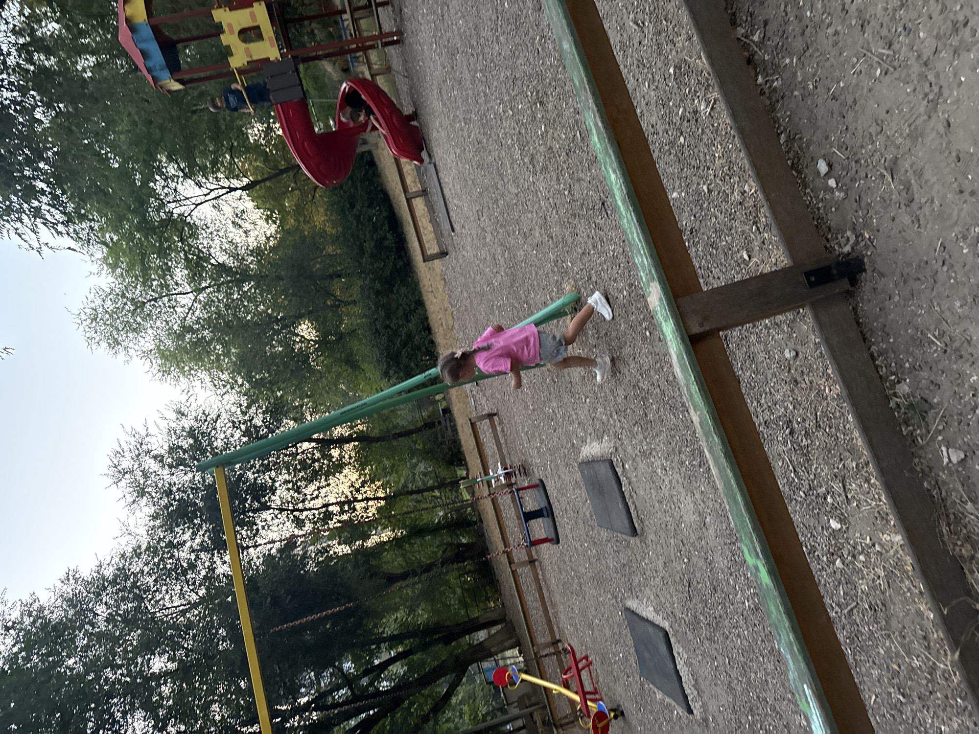 The image depicts an outdoor playground scene with several pieces of equipment. In the foreground, a young girl wearing a pink shirt and grey shorts is running on a gravel surface. A wooden fence with a green railing runs along the bottom of the frame. In the middle ground, there is a swing set with red and blue seats and a seesaw with red and yellow components. Further back, a red spiral slide is attached to a multi-colored play structure with yellow and red elements. Two children, one wearing a blue shirt and the other partially visible, are on or near the slide. The playground is surrounded by trees, suggesting a park or recreational area. The lighting suggests it is either early morning or late afternoon. The specific location in Chișinău, Moldova cannot be determined from the image alone.