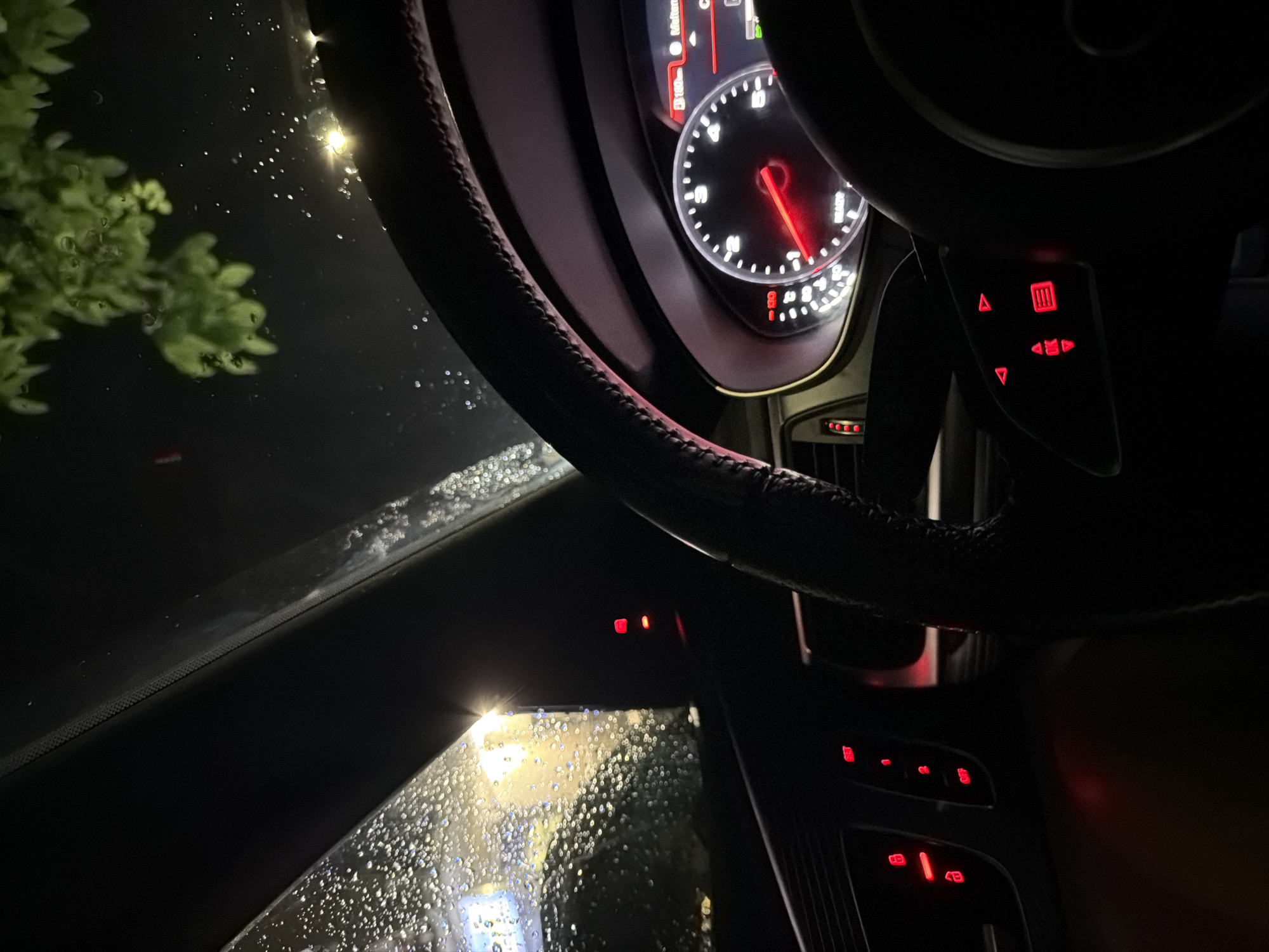 The image captures the interior of a car at night, likely in Veliko Tarnovo, Bulgaria, as indicated by the visual context. The focus is on the dashboard and steering wheel, illuminated by the soft glow of the car's instruments and the ambient light filtering in through the rain-streaked windshield. Water droplets are visible on the windshield, suggesting it is raining. Outside, the faint glow of streetlights or building lights can be seen, casting a yellowish hue on the wet glass. The dashboard displays a speedometer and other indicators, all lit with red and white illumination. The steering wheel, wrapped in dark leather, occupies a significant portion of the foreground. Control buttons on the steering column are also illuminated with red LEDs. The overall atmosphere is one of a quiet, introspective moment inside a vehicle during inclement weather. No people are visible in the image.