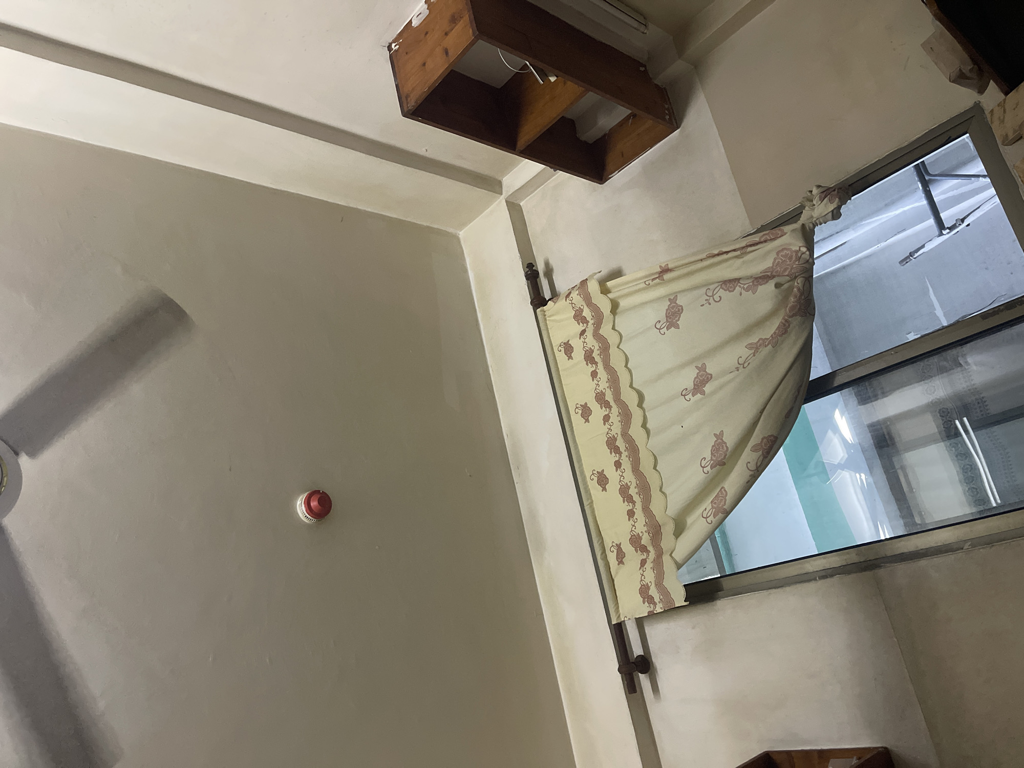 The image depicts an interior scene of a room, possibly in Dar es Salaam, Tanzania. The room is lit, and the walls are painted a light beige color. Visible on the ceiling is a light fixture, a ceiling fan (partially visible on the left), and a wooden shelf unit. A window is present, partially covered by a curtain with a floral pattern. A red circular object, possibly a smoke detector or a similar device, is attached to the wall. No people are visible in the frame.
