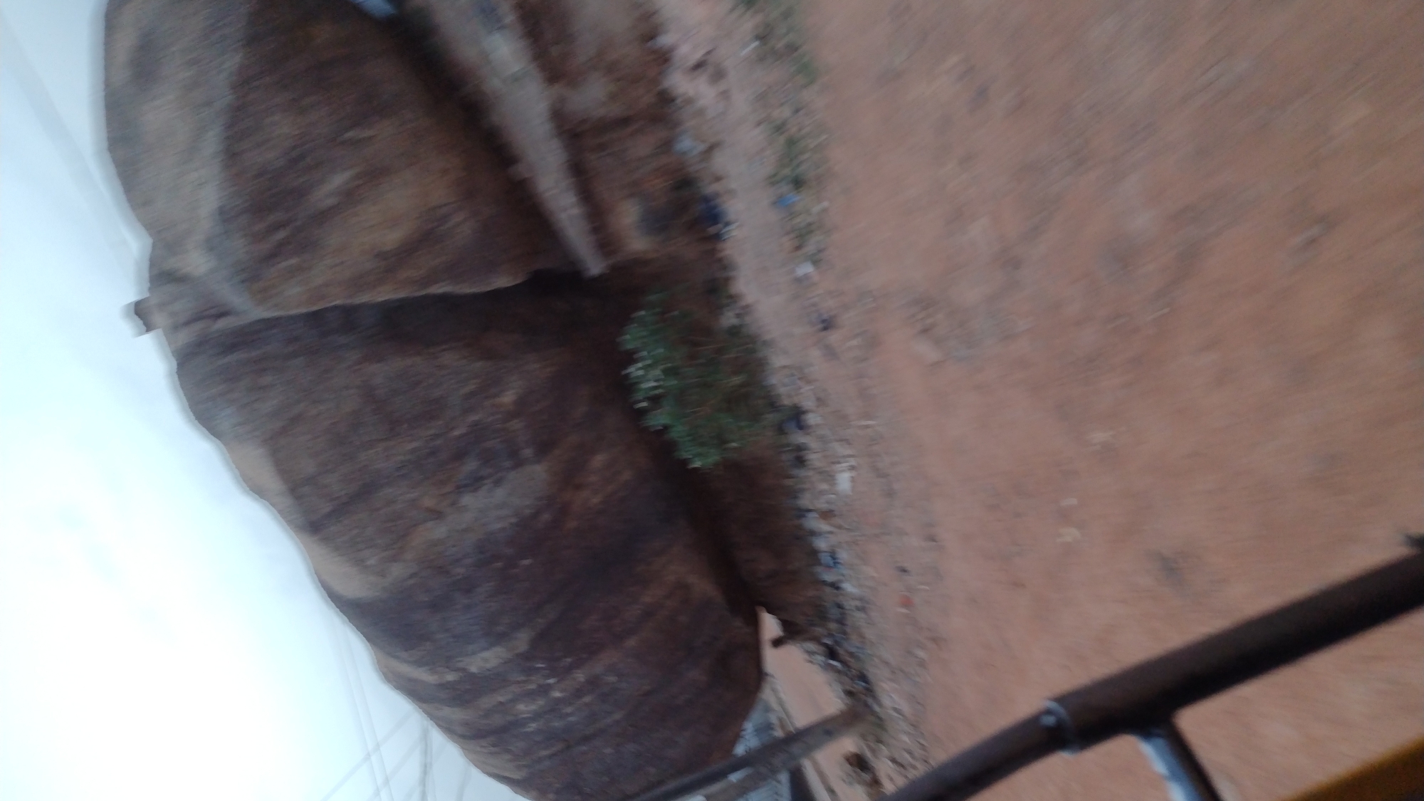The image depicts a rocky outcrop alongside an earthen surface, likely soil or dirt. Vegetation is sparsely present, growing near the rocks. Parts of a metal railing can be seen. The location is likely in or near Jos, Nigeria, due to the prevalence of such rock formations in the area. The absence of visible people suggests a natural environment or sparsely populated area.