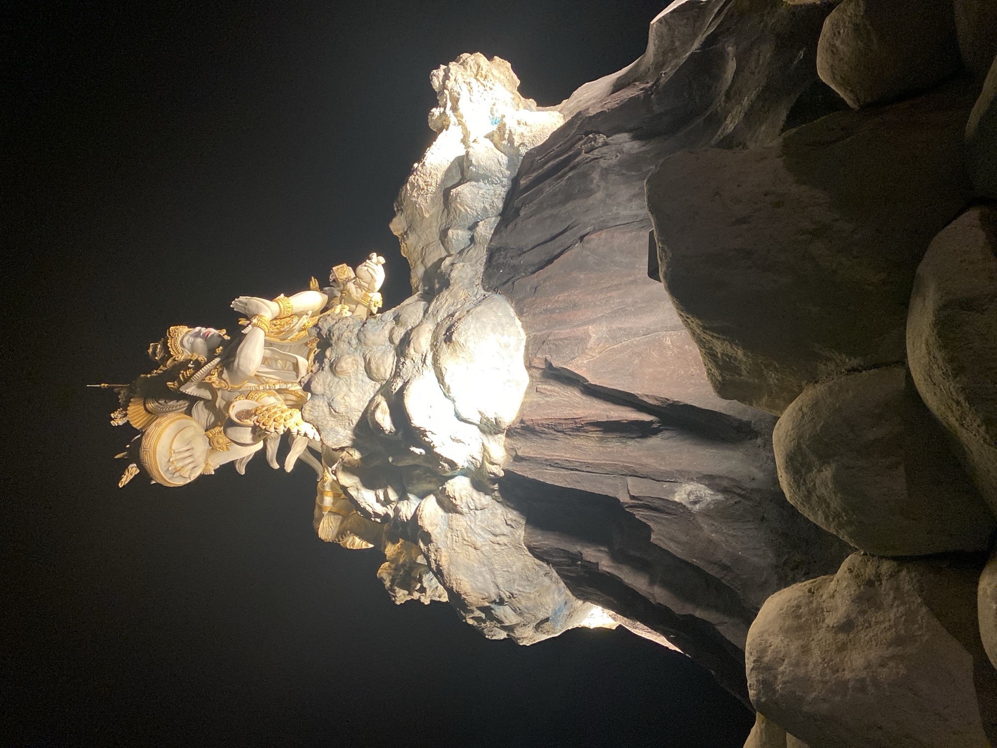 The image captures a nighttime scene in North Kuta, Indonesia, focusing on a religious sculpture or carving attached to a rock formation. A statue of Vishnu is carved into the rock formation. The statue is cream-colored with gold ornamentation. The sculpture is illuminated with a bright spotlight. The surrounding environment consists of dark rock formations and darkness. The image has been rotated counterclockwise.