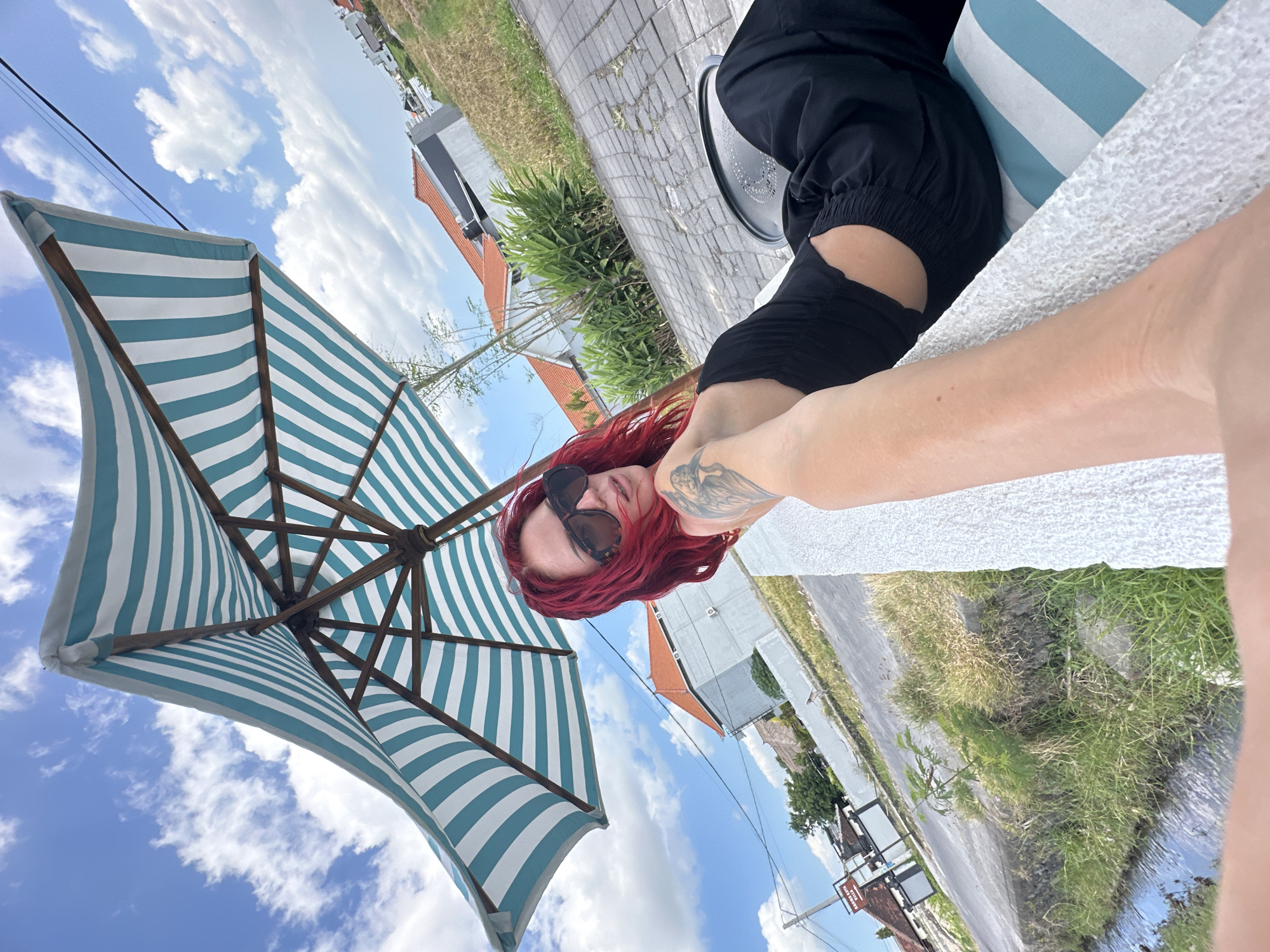 A woman with bright red hair and sunglasses is lying on a striped chaise lounge under a blue and white striped umbrella. She is wearing a black top and black pants. Her left arm is extended towards the camera, and she has tattoos on her arm. The chaise lounge is on a patio with brick pavers, next to a road with some greenery and buildings in the background. The sky is blue with white clouds. The location is indicated to be North Kuta, Indonesia, by signage in the background.