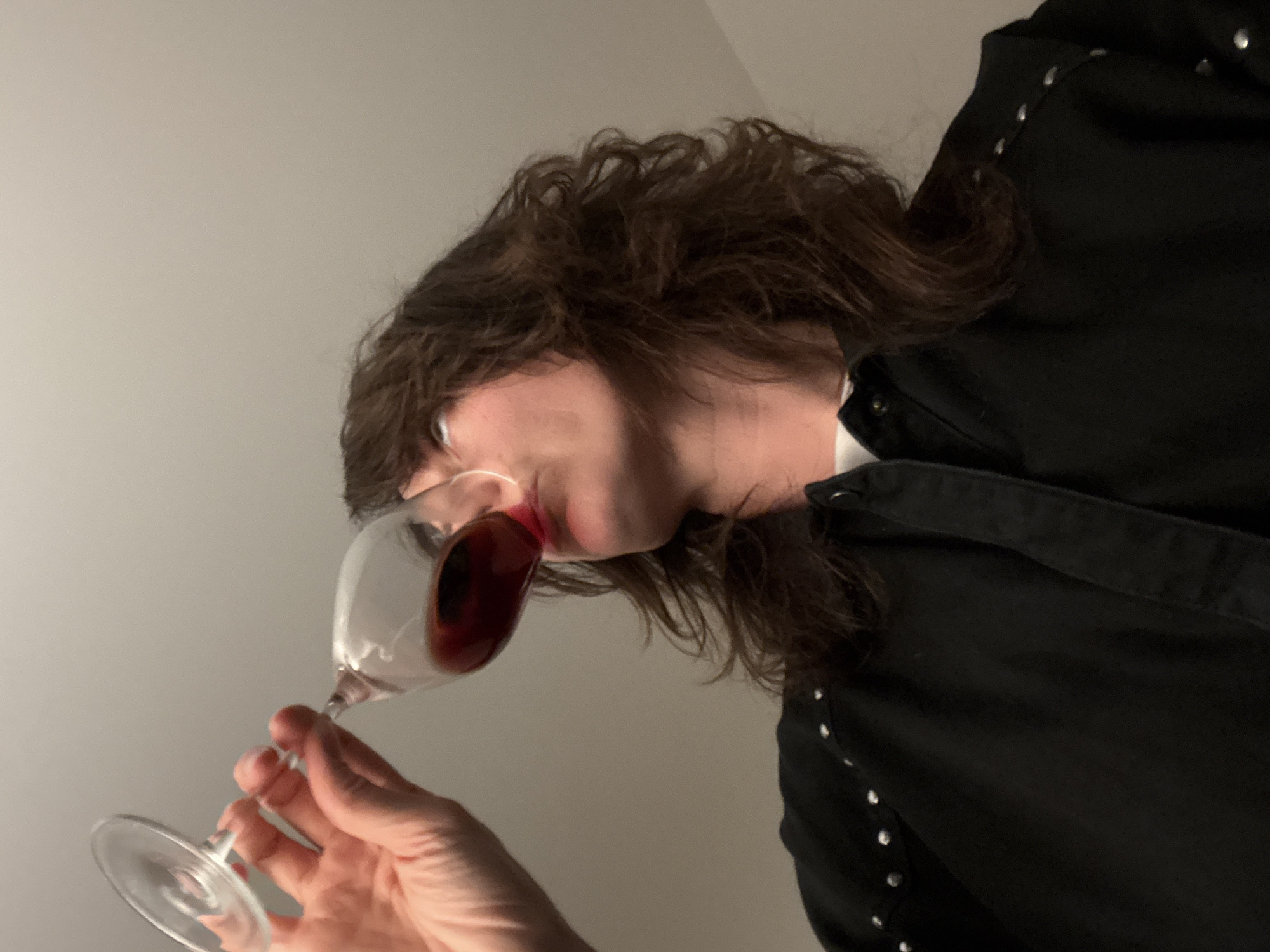 The image shows a person holding a wine glass filled with red wine, tilting it towards their mouth. The person has medium-length, wavy brown hair and is wearing a black, collared shirt with silver studs along the shoulder and sleeve. The background is a plain, light gray wall. No details suggest the location is in Boreti, Montenegro.