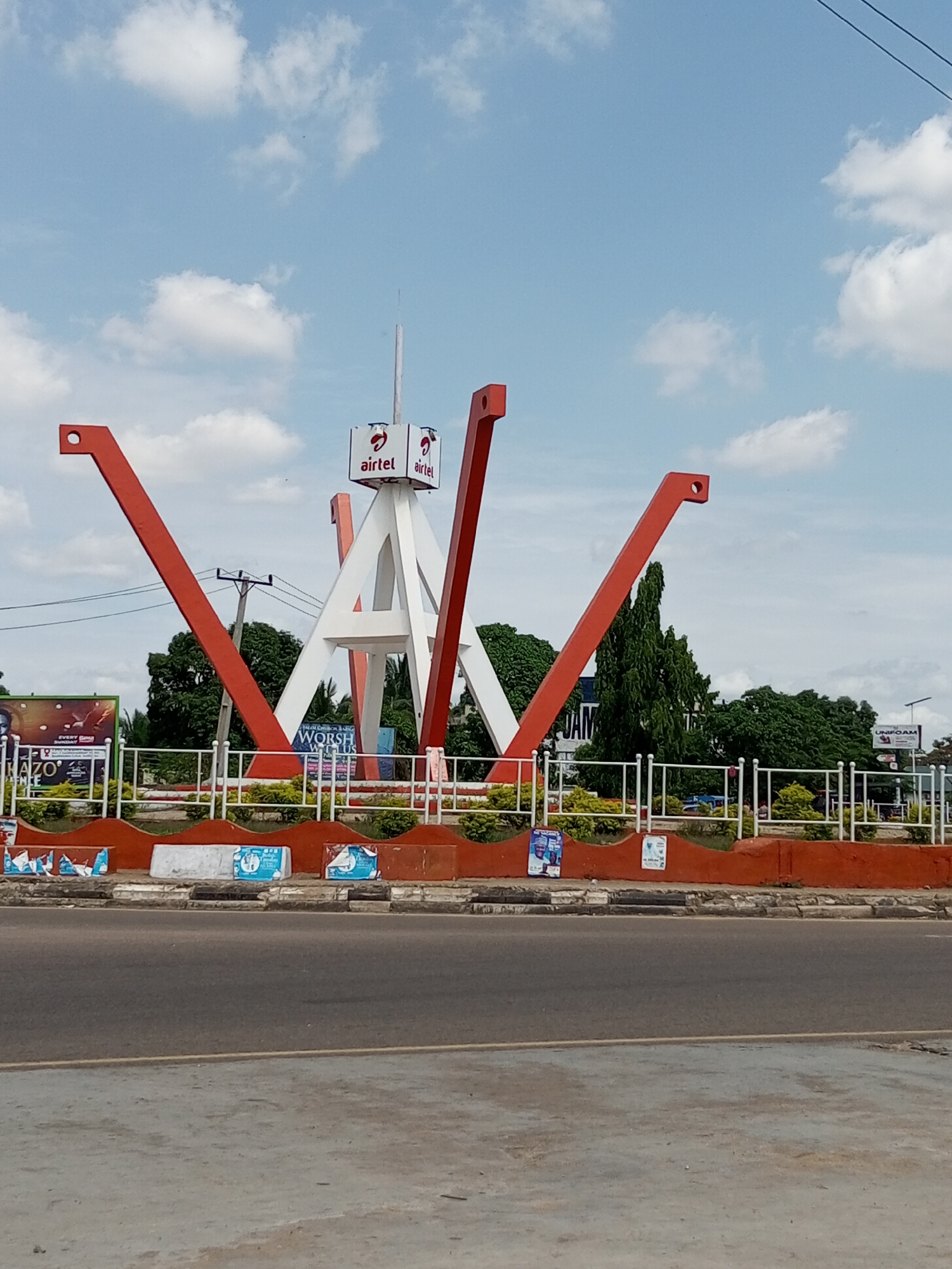 The image displays a daytime street scene in Barnawa, Nigeria, featuring a prominent red and white monument or sculpture situated within a landscaped central island. The sculpture comprises a central white structure, possibly forming a letter 'A' or triangular bases, from which multiple red, inverted V-shaped beams extend outwards and upwards, each culminating in a circular aperture at its apex. At the peak of the central white structure, a vertical pole supports three rectangular signs, each displaying the red and white "airtel" logo.
The central island is bordered by a low, curved red wall topped with white trim and a white metal railing. Green shrubs and small trees are planted within this area. Various advertisements and posters are affixed to the red wall and the ground. On the left, a large green and orange billboard is visible.
In the foreground, a dark grey asphalt road indicates a public thoroughfare, separated from the central island by a lighter grey concrete curb. Some discarded items or packaging are present on the curb area near the red wall. In the mid-ground behind the sculpture, green foliage from numerous trees is visible, along with utility poles and overhead power lines on the left. A partially visible billboard with "UNIFOAM" text can be seen on the far right. The sky overhead is bright blue with scattered white cumulus clouds. No people are discernible in the frame.