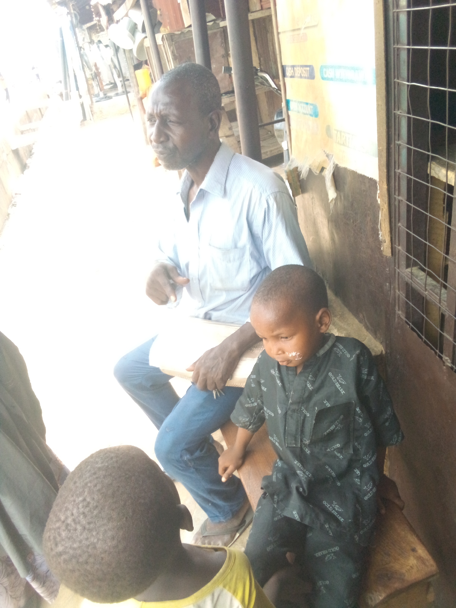 The image shows an adult male with closely-cropped, graying hair, wearing a light blue striped, button-down shirt and blue jeans. He sits on a wooden bench next to a young boy. The boy has a small amount of white substance, possibly food, on his face. He is wearing a dark patterned shirt and shorts, with "GOLD SHARK" visible on the fabric. The man is holding a white object, possibly a piece of paper or a small board. To the side, another child is partially visible, wearing a yellow shirt.
The background reveals an outdoor urban setting. A weathered wall with peeling signage is visible, with the words "CASH DEPOSIT", "CASH", and "ACCOUNT" partially readable. There is also a metal-barred window to the right. The overall lighting is bright, suggesting a sunny day. The location is likely an informal or commercial area, potentially in Minna, Nigeria.
