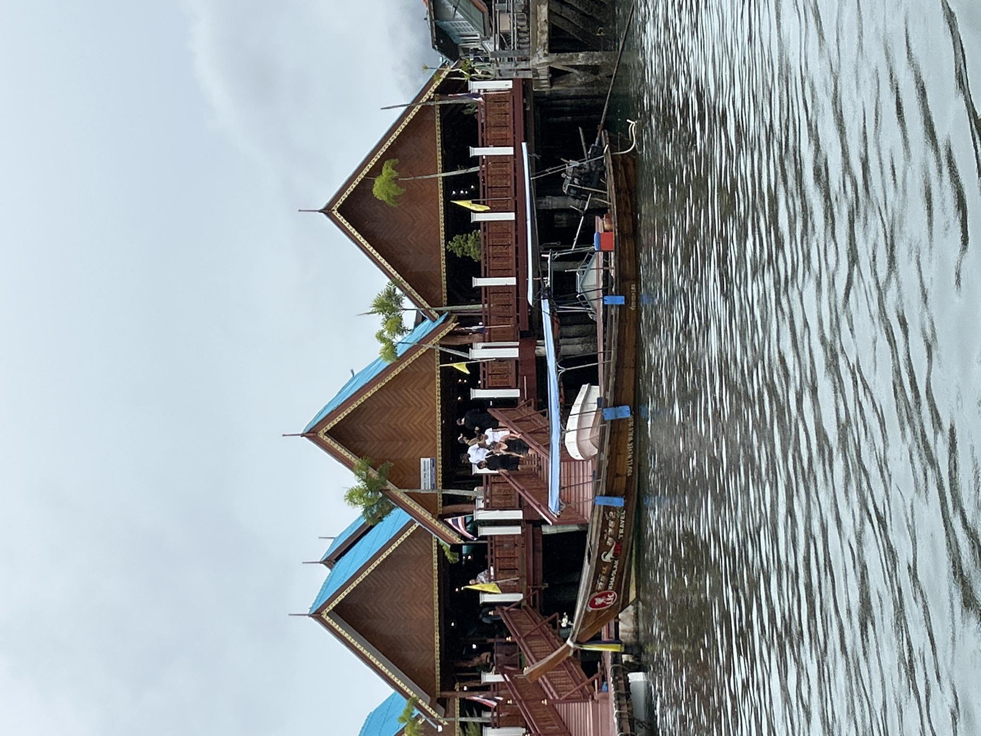 The image shows a view of the floating village of Ko Panyi, Thailand, from the water. In the foreground, the rippling surface of the water dominates the lower half of the frame. A long, narrow wooden boat, commonly known as a longtail boat, is moored alongside a wooden pier. The boat has a blue canopy and a small outboard motor at the stern. It features painted text that reads "LK KHAOLAK TRAVEL" and "YOU'LL NEVER WALK ALONE".
In the background, several wooden structures with distinctive steeply pitched, blue-tiled roofs rise above the water. These buildings are elevated on stilts, typical of the architecture in this stilted village. Red wooden stairs and walkways connect the buildings and lead down to the water. Several people are visible on the walkways and stairs, some appearing to be entering or leaving the buildings, while others are standing and observing. Yellow flags are displayed outside some of the buildings. The sky is overcast, suggesting a cloudy or possibly rainy day. The overall scene depicts a bustling, waterfront community.