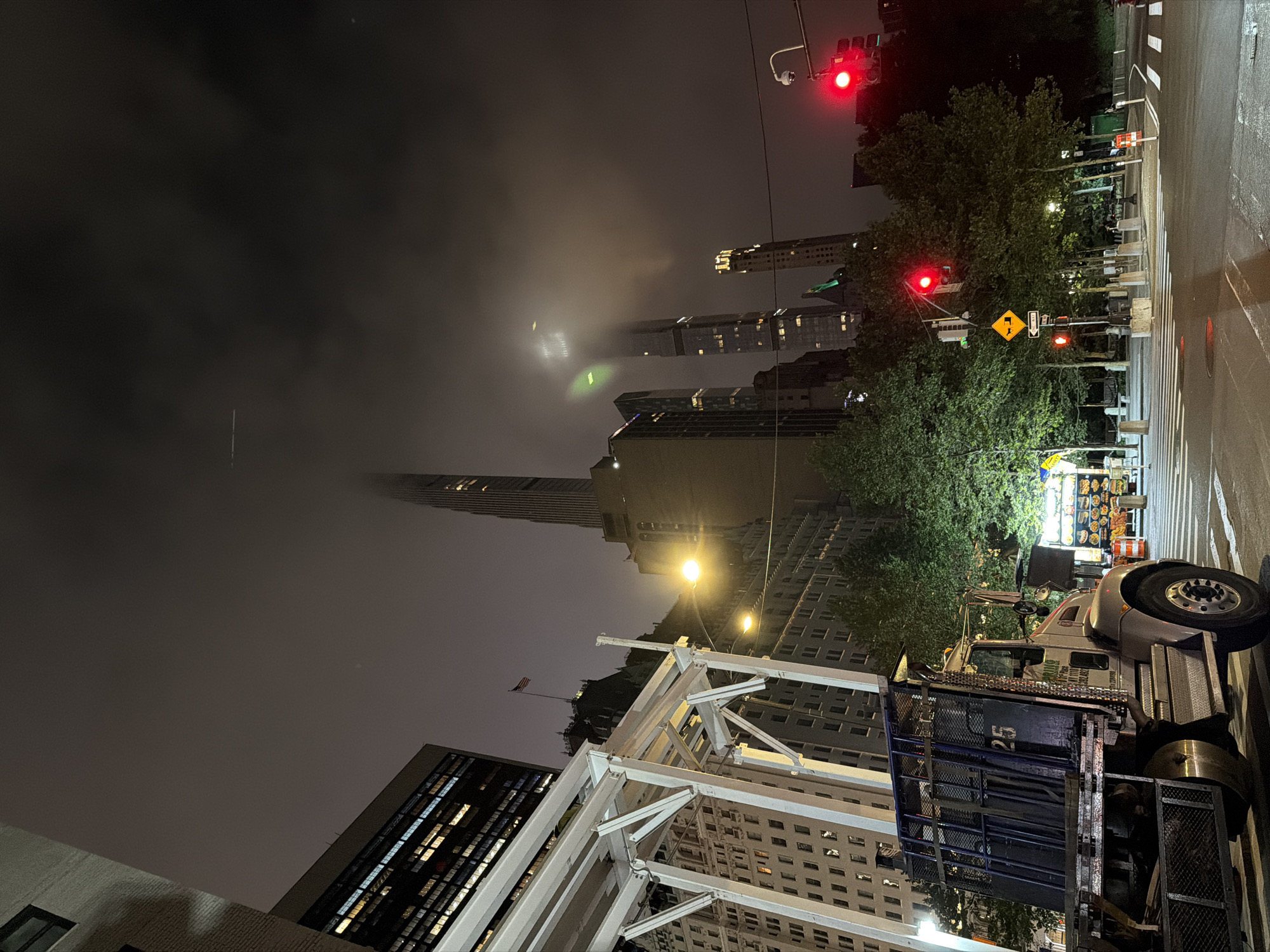 An eye-level, full shot captures a dimly lit, cloudy night scene in New York City. The foreground is dominated by a parked truck, its metallic blue cage visible on the rear. To the left, a white, modern architectural structure with angled supports stands out against a backdrop of older, ornate buildings. The midground features trees illuminated by streetlights, with a street vendor's cart displaying various foods. The background is a cityscape of towering skyscrapers, some obscured by fog, with a faint airplane contrail visible in the dark sky. Traffic lights display red, and a yellow diamond-shaped road sign with a silhouette of a vehicle is present.