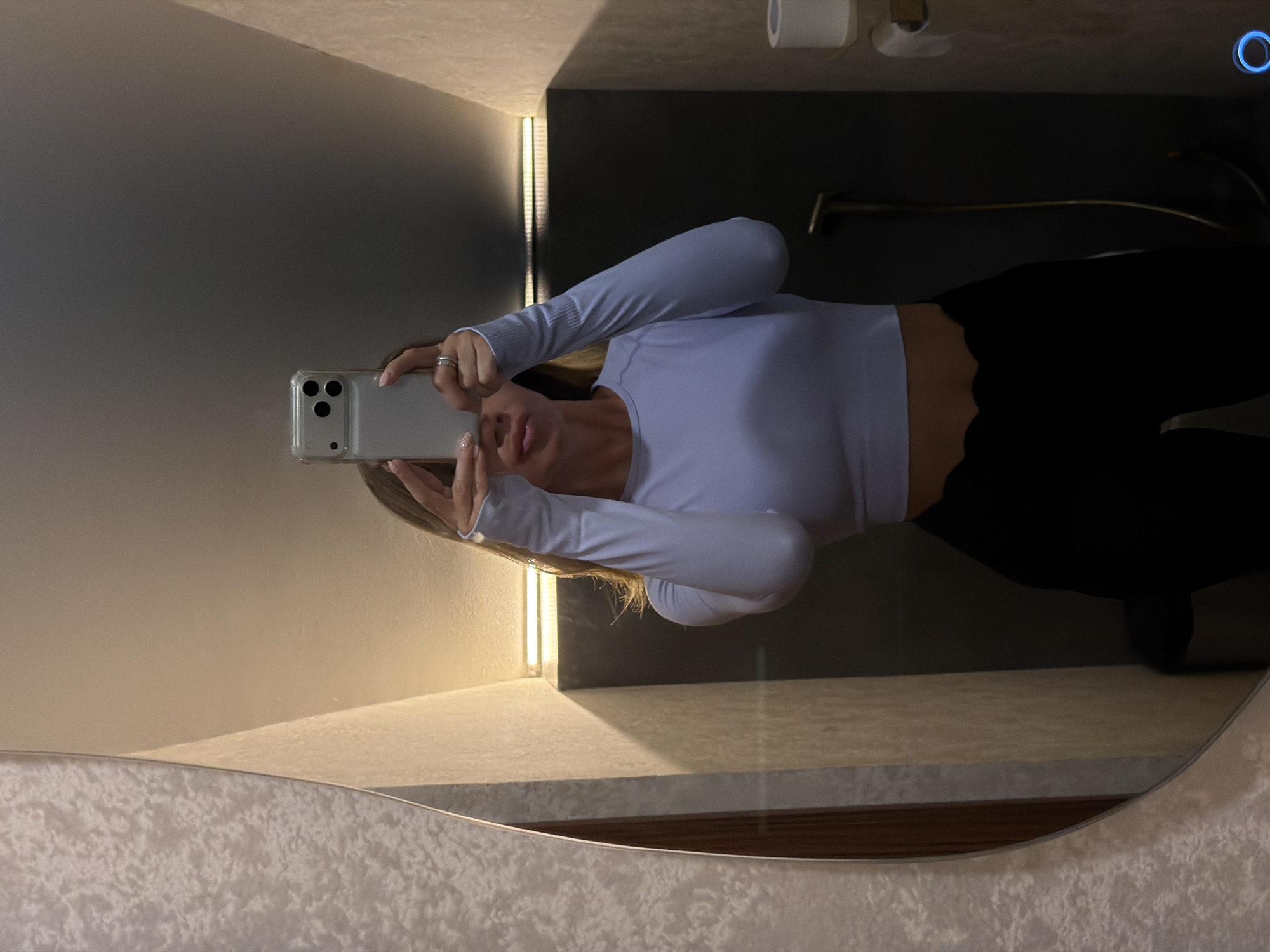 The image is a mirror selfie taken in a bathroom. The subject is a person holding a smartphone and taking a photo of themselves. They are wearing a light blue long-sleeved top and black pants. The background of the bathroom is visible, including a toilet paper holder, a faucet, and a light fixture. The overall impression is that of a casual, everyday moment. The lighting suggests it might be daytime or the lights are on in the bathroom. The reflection in the mirror shows the person's face and torso. The person is looking at the camera and appears to be posing for the selfie. The bathroom decor is modern and clean, with a textured wallpaper on one wall. The image is taken from a slightly high angle, looking down at the person.