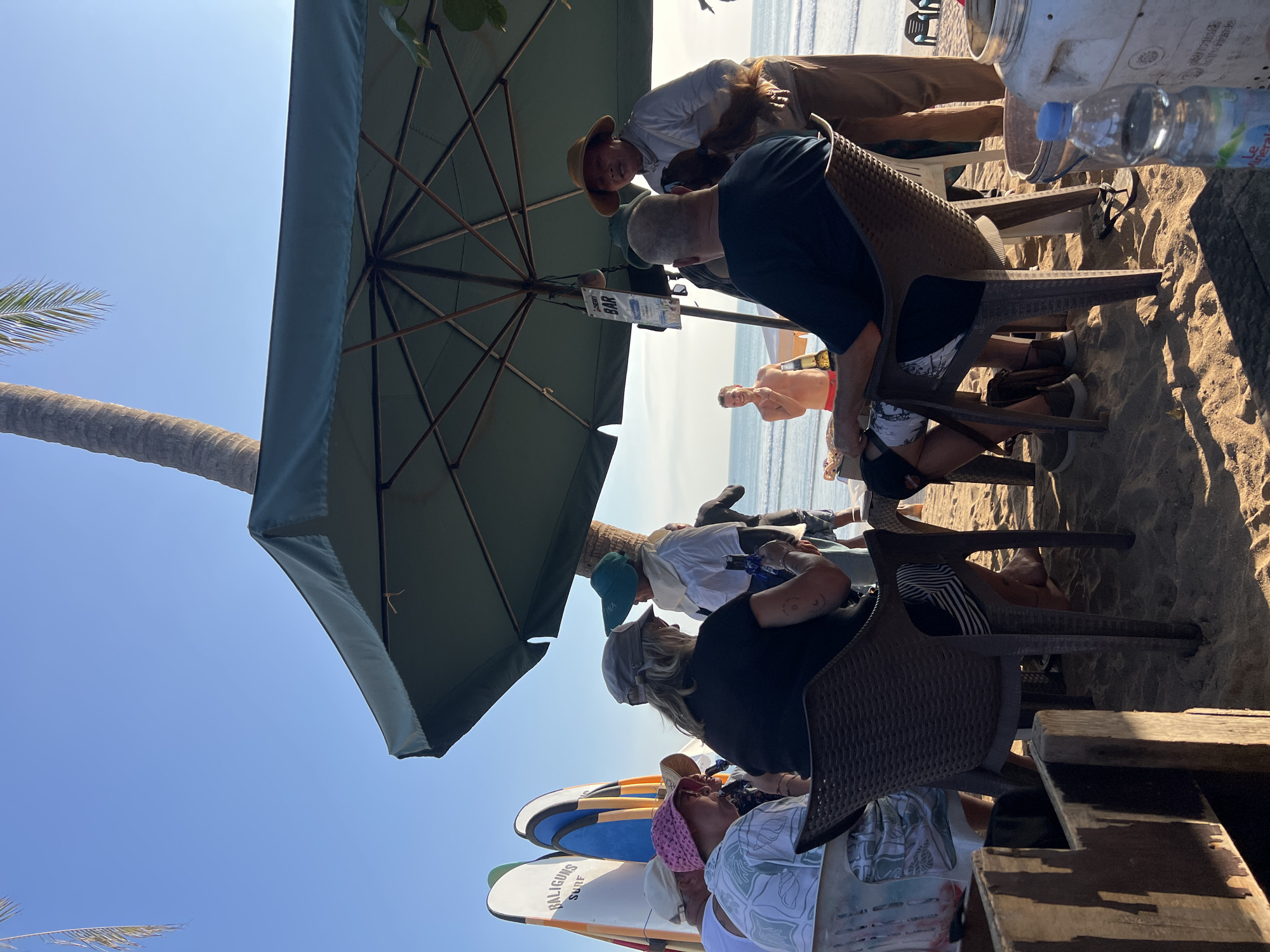 The image depicts a beach scene in Kuta, Indonesia, under a bright, clear sky. Several people are gathered around tables and chairs set up on the sand, likely at an outdoor cafe or bar. A large green umbrella provides shade.
In the foreground, a woman wearing a pink hat and a patterned shirt is seated at a table, facing away from the camera. To her left, another person in a grey cap and black shirt is also seated. A dark-colored plastic chair is visible in the mid-ground, occupied by a person wearing a dark blue shirt and patterned shorts.
Behind the seated individuals, a man in a straw hat and light-colored clothing appears to be interacting with someone, holding what looks like a brown object. To the left, several surfboards with "BALIGUNS SURF" branding are propped up.
In the background, the ocean is visible with gentle waves, and a few beach chairs are placed further down the sand. A man in red shorts is standing on the beach, holding a bottle. The environment is sandy, with shadows cast by the umbrella and surrounding objects. A sign on a pole near the umbrella reads "CHERRY BAR." The overall impression is a relaxed beach atmosphere.
