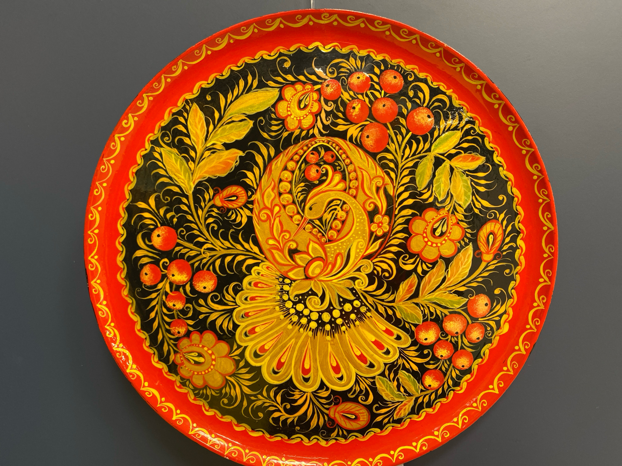 A decorative, circular tray is displayed against a dark gray wall. The tray features a vibrant Khokhloma painting, a traditional Russian folk art style. The design is predominantly black, red, and gold. In the center, a stylized golden bird with intricate feather details is depicted, surrounded by floral and berry motifs. The outer edge of the tray is bright red with a decorative golden border. The image shows no people or animals. The setting appears to be an indoor exhibition or display space. The specific location in Ivanteyevka, Russia, cannot be determined from the image alone.