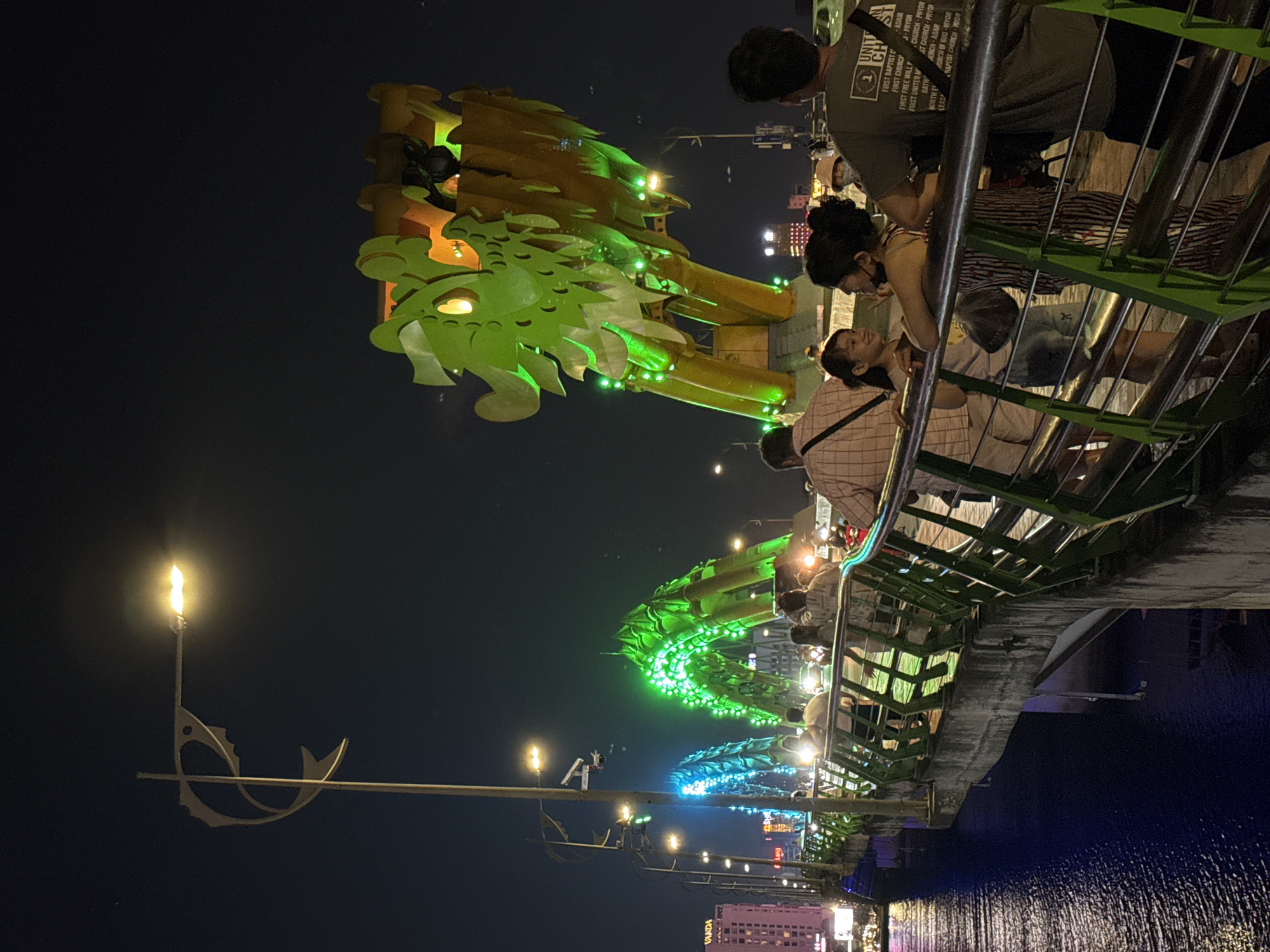 The image presents a vibrant night scene on the Dragon Bridge in Đà Nẵng, Vietnam. The central feature is a large, illuminated dragon sculpture, predominantly green and gold, stretching across the frame. Its head, adorned with bright green lights, is prominent on the left, with the body extending towards the right.
Several people are visible on the bridge's pedestrian walkway, which features a green railing. In the foreground on the right, a person in a dark green t-shirt with extensive white text stands with their back mostly to the viewer, observing the scene. Beside them, two other individuals, one in a striped top and another in a plaid shirt, are engaged in conversation, appearing relaxed. Other people are scattered along the bridge, enjoying the evening.
Below the bridge, dark water reflects the city lights, creating shimmering patterns. On the far left, a distinctive street light in the shape of a fish, topped with a glowing yellow-orange lamp, adds to the unique urban aesthetic. In the distance across the water, various buildings are visible with their lights on, including one with the illuminated name "VANDA."
The activity depicts people leisurely strolling and observing the famous Dragon Bridge and its surroundings at night, suggesting a popular tourist spot or local attraction. The time of day is clearly night, as indicated by the dark sky and numerous artificial lights. The atmosphere appears calm and enjoyable.
Visible text includes:
On the dark green t-shirt: "UNIT CH", "FIRST BAPTIST ADAIR", "FIRST CHURCH", "FIRST FREE WILL P", "J-PRYOR", "CHURCH-PRYOR", "FIRST UNITED MST CHURCH-PRYOR", "GATH", "NORTH E", "LEAS", "HURCH-ADAIR", "HURCH OF GOD-PEYOR", "RAPTIST CHURCH-PRYOR".
On a distant building: "VANDA".