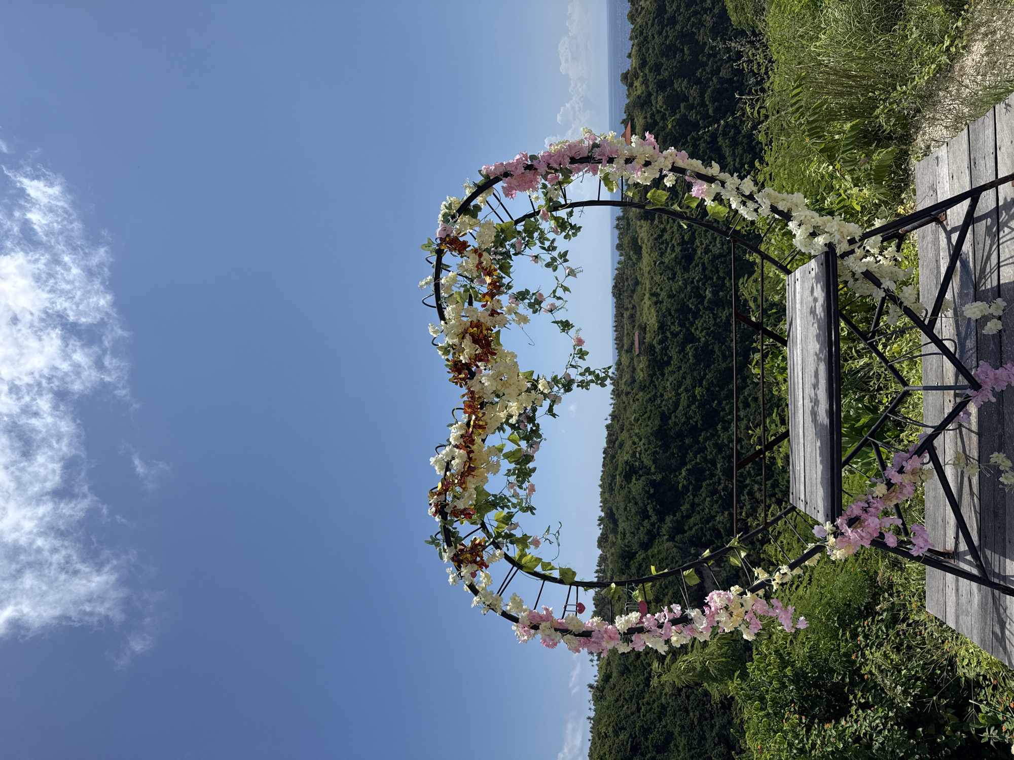The image captures a heart-shaped arch decorated with pink, white, and orange flowers. The arch is positioned on a wooden platform overlooking a dense green forest. In the background, a body of water can be seen separating the forest from the sky. The sky is bright blue with some scattered clouds. The location is Pejukutan, Indonesia.