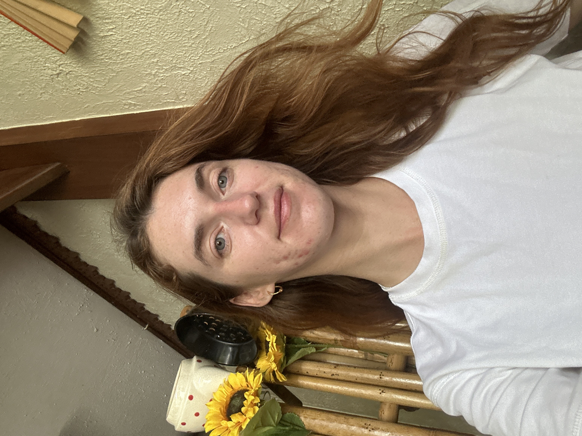 A woman with reddish-brown hair lies on a bamboo bench, wearing a white t-shirt. She is fair-skinned, with light eyes and some facial blemishes. Near her head are yellow sunflowers and a black object, possibly a diffuser. In the background, there's a cream-colored wall, a dark wood trim, and a stack of books. The setting is suggested to be in Baan Chaweng Noi, Thailand.