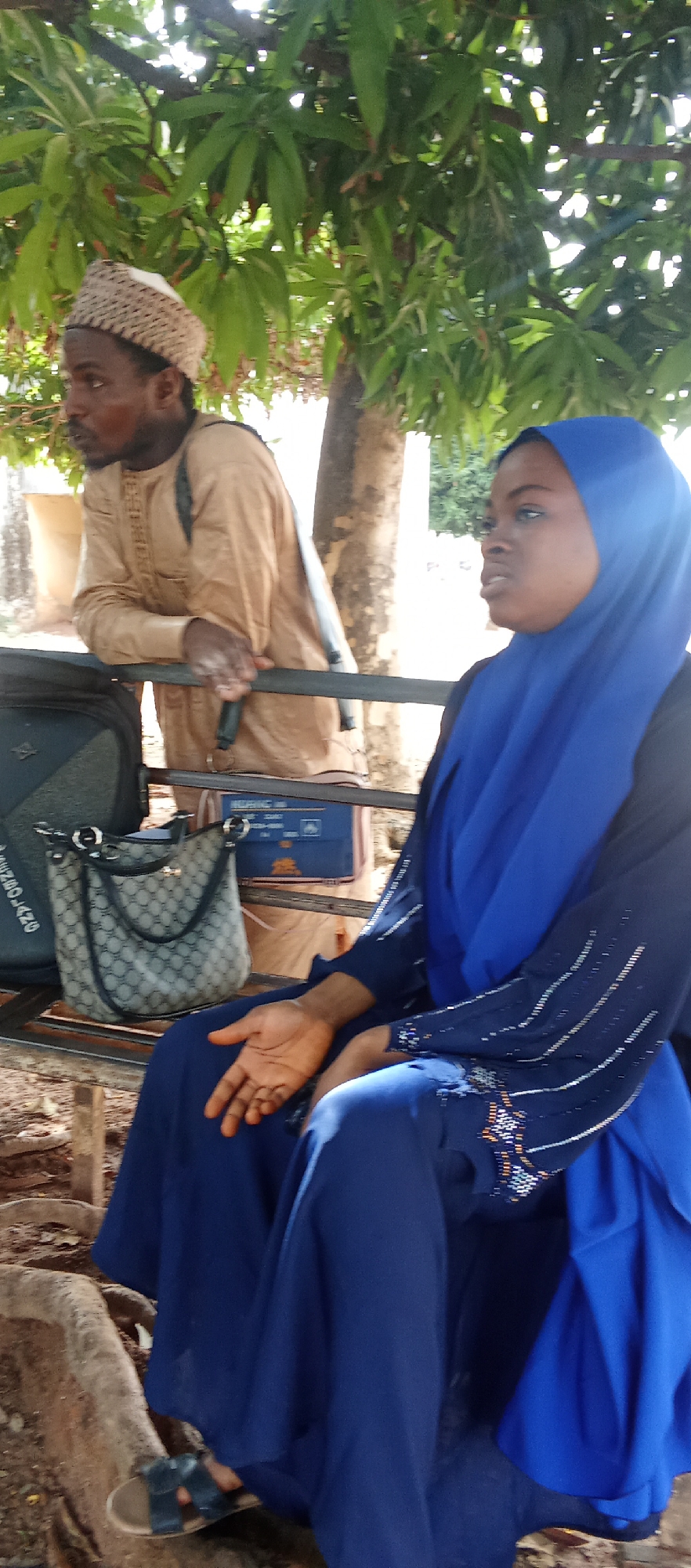 The image shows a man and a woman in what appears to be an outdoor setting. The man is standing behind a metal structure, wearing a light brown traditional outfit and a woven cap. He has a backpack and is looking off to the left. The woman is seated, dressed in a long blue gown and hijab. She is also looking to the left and gesturing with her right hand. Next to her is a handbag and a suitcase. The background includes green trees and a building in the distance. The location is specified as Akwanga, Nigeria.