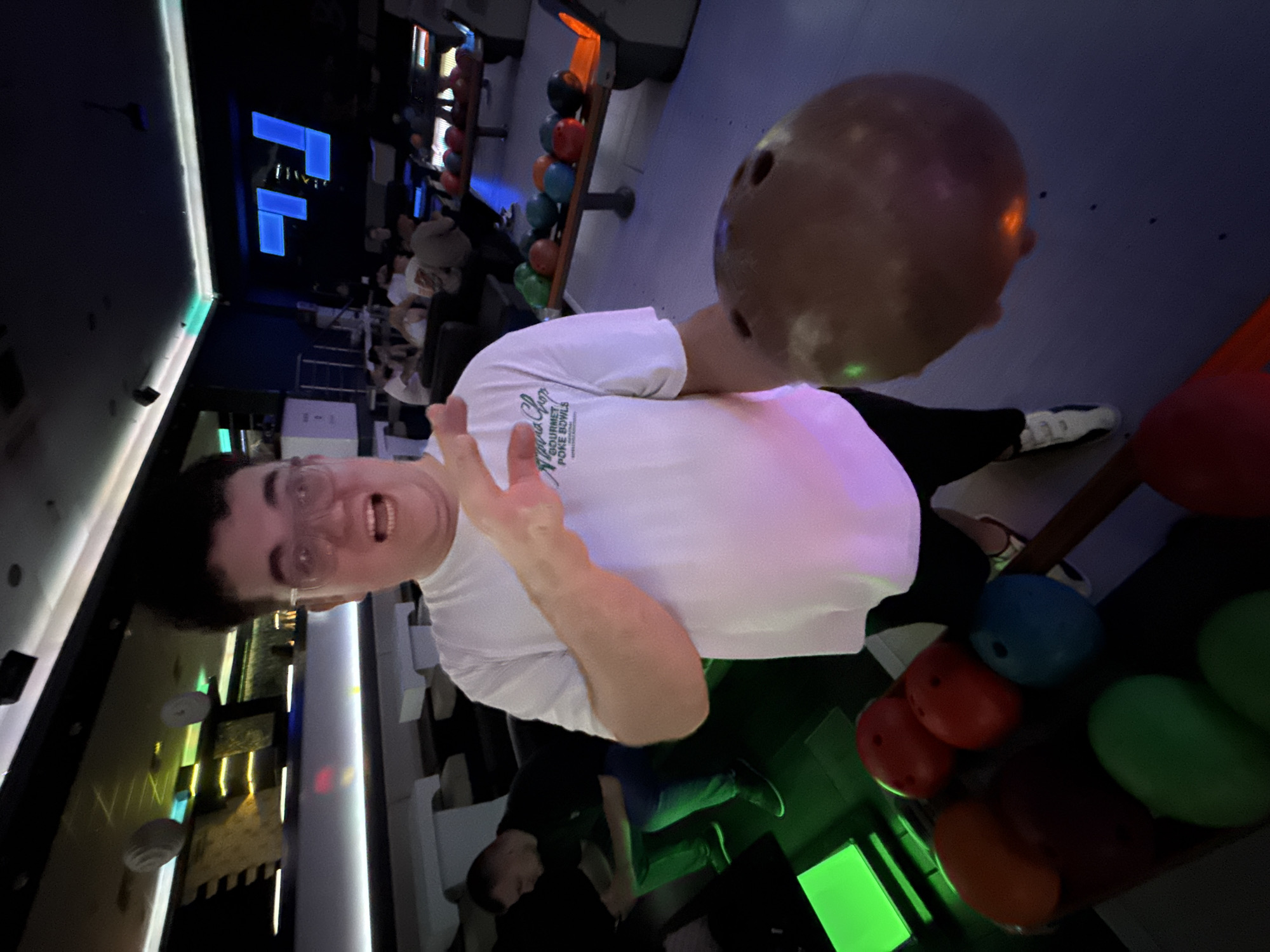 The image shows a man in a bowling alley, holding a bowling ball and making a peace sign with his hand. He is wearing a white t-shirt with a logo that says "Napua Chop Gourmet Poke Bowls." Behind him, there are other bowling balls of different colors, and people can be seen in the background. The location appears to be a bowling alley, but the specific city of Pleven, Bulgaria, cannot be definitively confirmed from the visual information alone.