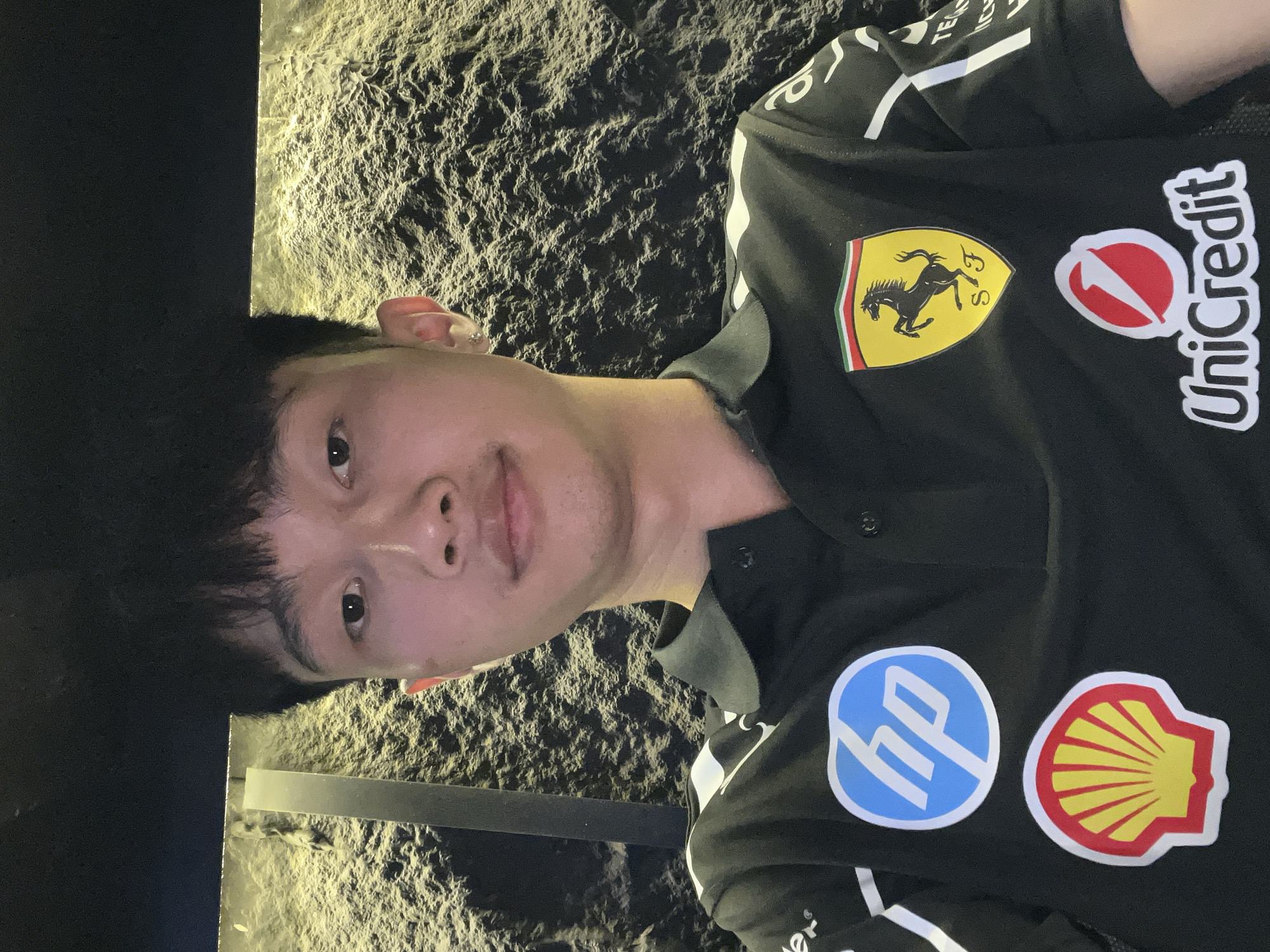 A young East Asian man is depicted in a close-up, chest-up shot, looking directly at the camera with a neutral expression. The image is vertically oriented, causing the man to appear horizontally in the frame. He has short, dark hair and fair skin.
He is wearing a black polo shirt, possibly a team or fan jersey, adorned with several prominent sponsor logos. On the upper left side of his chest, the yellow shield logo of Ferrari is visible, featuring the black prancing horse, "SF" initials, and the green, white, and red stripes of the Italian flag. Next to it is the UniCredit logo, a red arrow within a circle alongside the "UniCredit" text. On the lower right side of the shirt, a blue circular logo with white "hp" text for HP is displayed, along with the distinctive yellow and red seashell logo of Shell. White stripes and some indistinct white text are also visible on the shirt's sleeves and shoulders.
The background features a dark, highly textured wall or surface, possibly rough stone or concrete, illuminated by a warm, yellow light source that creates strong highlights and shadows across its uneven topography. Hints of metallic or reflective strips, potentially part of a railing or architectural detail, are also visible. The dim and focused lighting suggests an indoor setting or an outdoor nighttime environment, consistent with a modern urban location in Đà Nẵng, Vietnam.