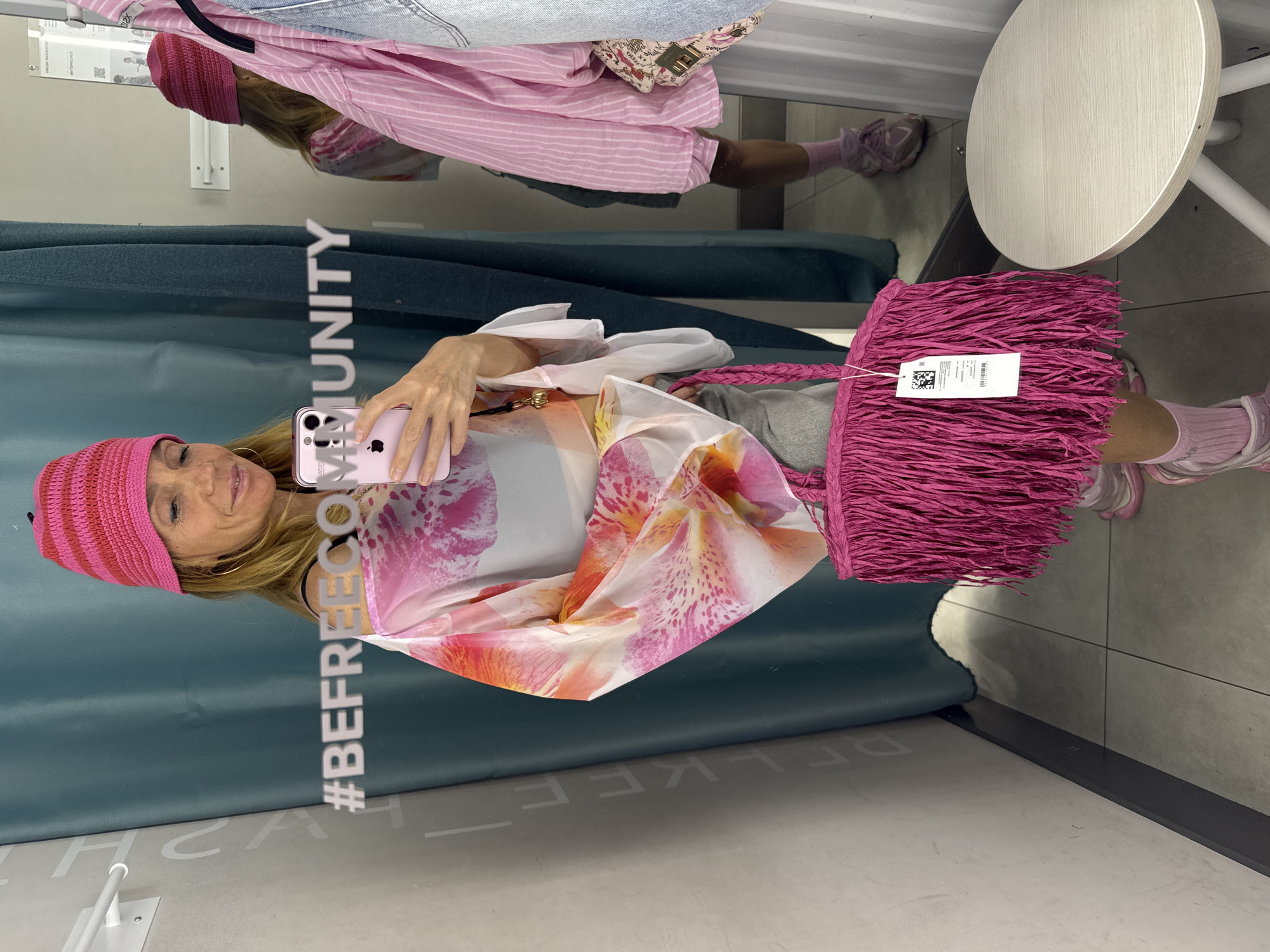 A woman is standing in a changing room, taking a mirror selfie. She is wearing a pink and red striped knit hat, a sheer pink and white floral print top, gray shorts, pink socks, and pink sneakers. She is holding a large pink fringed bag and a pink smartphone. The changing room has teal curtains and a white stool. A hashtag "#BEFREECOMMUNITY" is visible on the mirror. The image is taken in Vsevolozhsk, Russia.