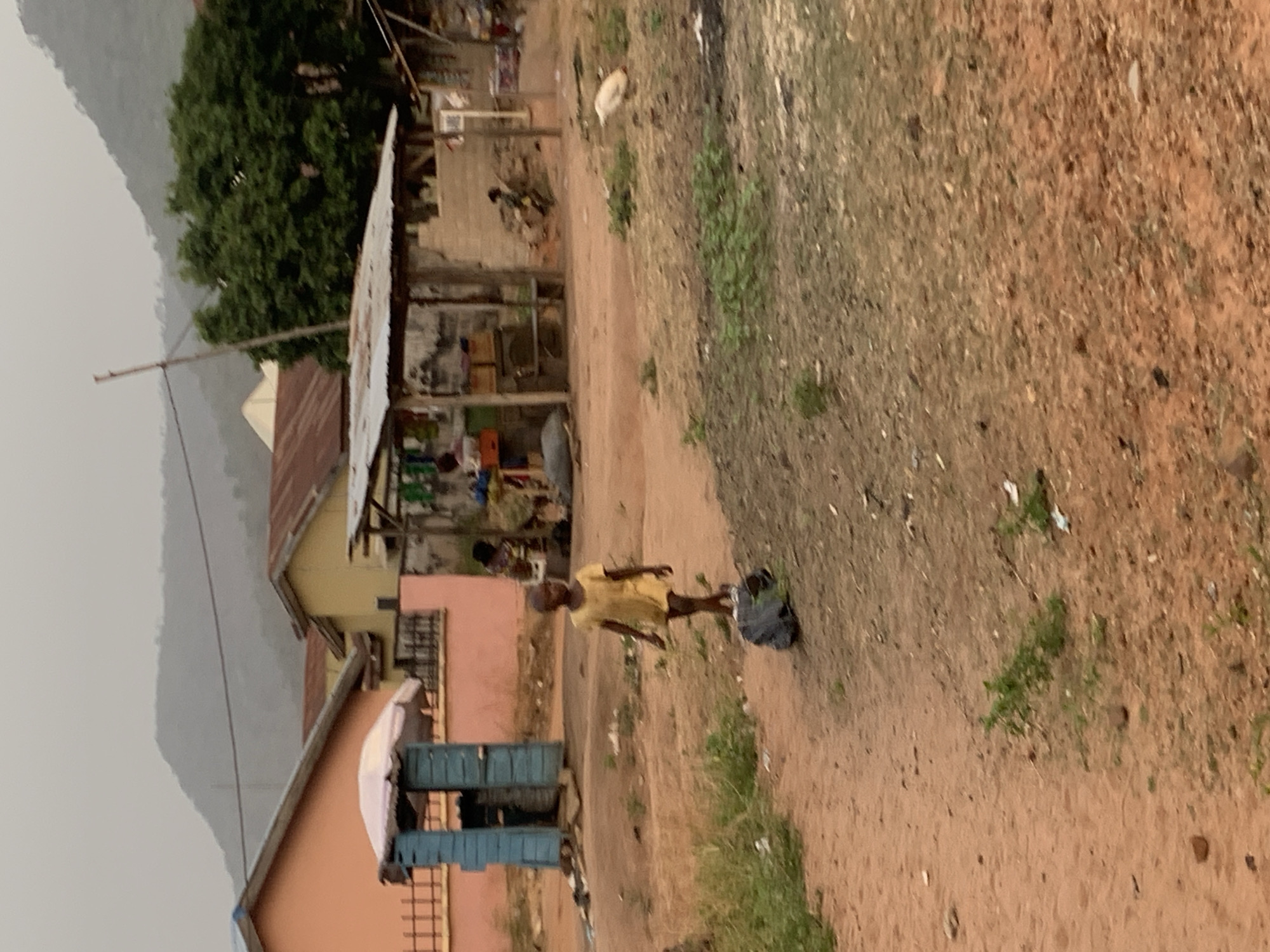 The image depicts a scene in Lokoja, Nigeria, likely a rural or less developed area. A young person wearing a yellow shirt and shorts is walking on a dirt path. A black bag is near them on the ground. Several simple structures with corrugated metal roofs and partially constructed walls, possibly houses or shops, line the side of the path. One structure appears to be a small store, with items visible on shelves. Other houses with red-tiled roofs are also visible. To the left, a mountain is discernible in the distance, with overcast weather. The ground is dirt and dry grass, showing sparse vegetation. A white chicken is present behind one of the shops.