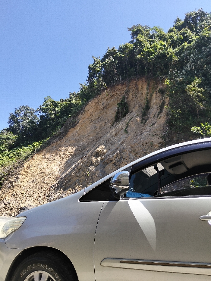 A silver car sits against a backdrop of a steep, eroded hillside, topped with dense green trees. The earth is a mix of pale browns and tans, showing signs of past landslides. The blue sky is visible above the trees and to the side of the hillside. The car’s tire, headlight, mirror, door, and tinted window are visible in detail.