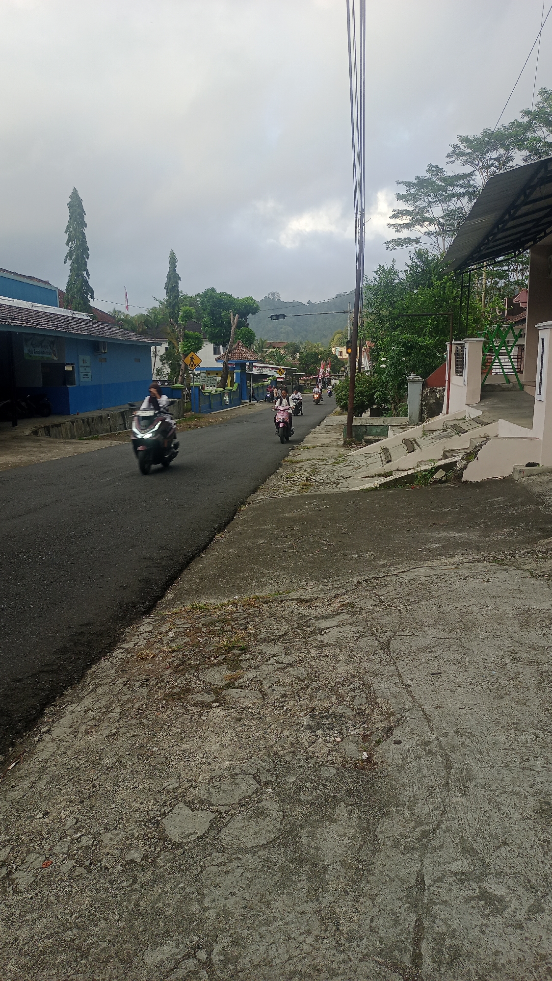 The image depicts a street scene in Karyamandala, Indonesia, under an overcast sky. The road is paved with asphalt and shows signs of wear. On the left side of the road, a blue building with a white trim is visible, possibly a public facility, with a flagpole displaying a Indonesian flag. Several trees, including tall, slender ones, are present along the road and in the background. A cluster of buildings with tiled roofs lines the right side of the street, some featuring awnings.
Several individuals are riding motorcycles on the road. One motorcycle in the foreground, a dark-colored scooter, is brightly lit by its headlights and appears to be in motion, with its rider visible. Further down the road, other motorcycles with riders are also traveling, with some appearing closer together, suggesting a small procession or group. Traffic signals are visible in the distance. The ground in the foreground is a concrete surface, cracked and weathered, with patches of dirt and sparse vegetation. Overhead, electrical wires crisscross the sky, supported by poles. The overall impression is a quiet, everyday scene in a semi-urban or rural Indonesian setting.