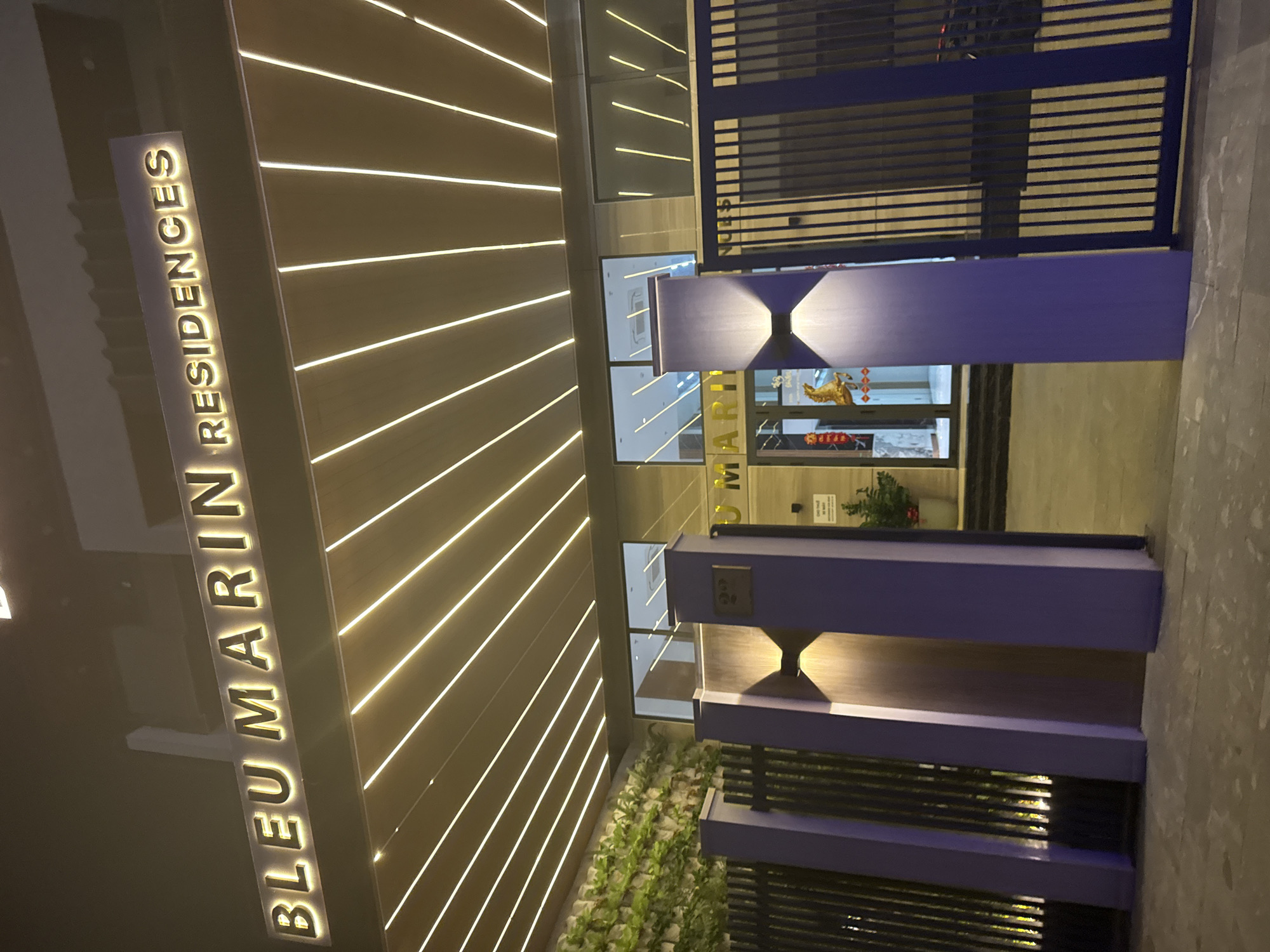 This nighttime image captures the illuminated entrance of the "BLEU MARIN RESIDENCES" building in Đà Nẵng, Vietnam. A prominent vertical sign on the left side of the frame displays "BLEU MARIN RESIDENCES" in glowing, capital letters. The building's facade to the left features a pattern of bright horizontal strip lights, creating a modern and dynamic aesthetic.
The central entrance consists of glass doors, through which a golden ox figurine and some potted plants are visible, hinting at a reception or lobby area inside. Framing this entrance are two sturdy, purple-colored pillars, each equipped with upward and downward-facing integrated light fixtures that cast a soft glow. To the right of the pillars, a dark blue metal gate with horizontal slats appears to secure the perimeter. A small garden bed with lush green plants is situated at the base of the left facade, adding natural elements to the contemporary design. The ground visible in the foreground is a grey tiled or concrete surface. The setting appears to be an urban environment, emphasizing the building's modern architecture and welcoming, yet secure, atmosphere. No people or specific activities are present. The distinct text "BLEU MARIN" is also partially visible on the building's facade, reflected or printed near the entrance.