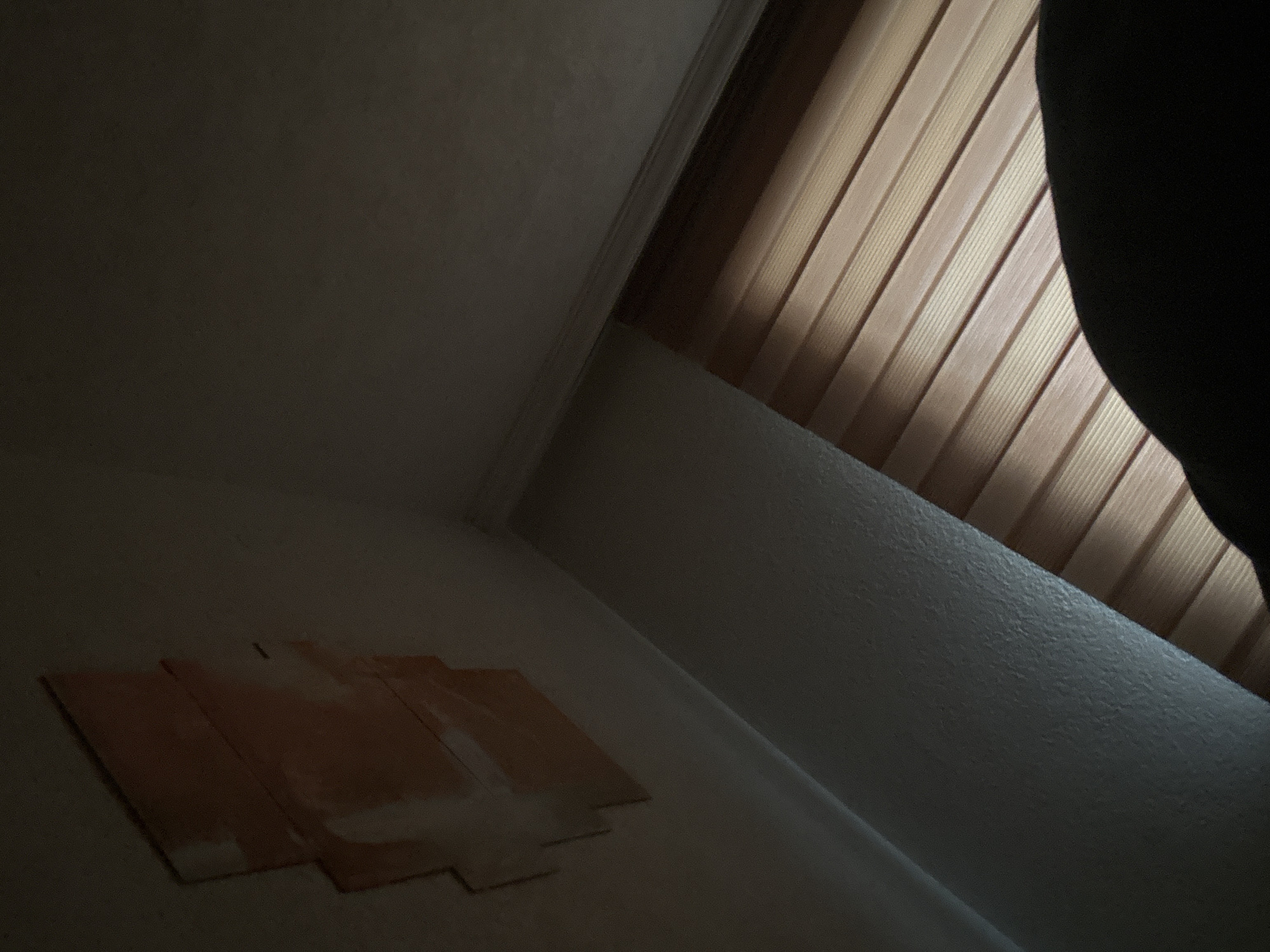 An interior scene within a room in Zlatograd, Bulgaria, is depicted under dim lighting. The upper-left quadrant of the frame shows a light-colored, textured wall to which four rectangular, orange-brown patches are affixed in a staggered vertical alignment. Apparent residue or lighter marks surround these patches on the wall surface. The upper-right corner features a similar light-colored, textured ceiling intersecting with a wall, separated by a distinct white crown molding or trim. A window, partially visible on the right side of the image, is fitted with horizontal zebra blinds exhibiting alternating stripes of translucent and opaque light beige and brown material, allowing light to filter into the room. A large, indistinct, curved dark mass occupies the bottom portion of the frame, obscuring the lower part of the window and blinds. No individuals are clearly identifiable, and no discernible actions or interactions are occurring.