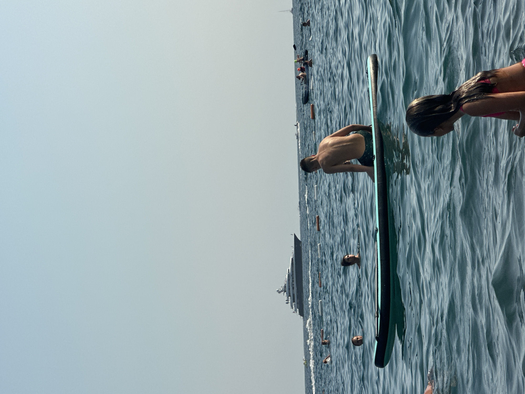 The image shows a group of people in the water at Boreti, Montenegro. In the foreground, a person on a paddleboard is seen from behind, with their torso exposed. To the right, another person, a child, is also on the paddleboard, with their back to the camera and their head bowed. The water is calm with gentle waves, suggesting a pleasant day. The sky is clear and pale blue, indicating daytime with fair weather. In the background, a large ship is visible in the distance, and several other people are scattered throughout the water, some of whom appear to be swimming or playing. The overall scene evokes a relaxed, summer atmosphere with people enjoying a day at the beach.