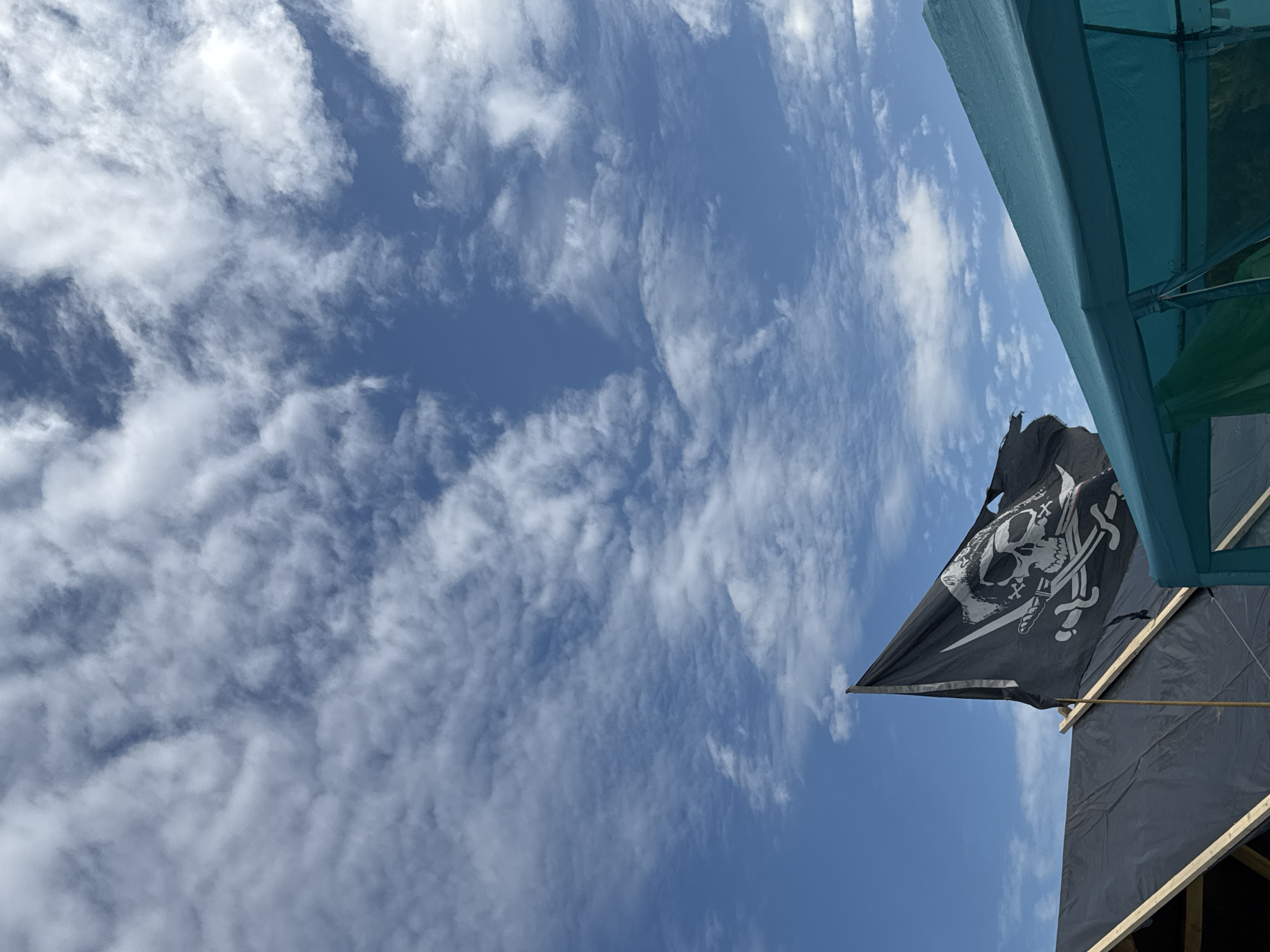 The image displays an outdoor scene in Большая Кивара, Russia, under a blue sky populated by scattered white cumulus clouds. In the lower-middle left, a black pirate flag, emblazoned with a white skull and crossed sabers (Jolly Roger), is visible, flying from a pole. The flag shows signs of wear, exhibiting tears and frayed edges. Beneath the flag, a structure with a pitched roof is covered by a dark, possibly black, tarp, with light-colored wooden beams exposed beneath. In the lower-right corner, a section of a green-teal colored tent or canopy is present, featuring taut fabric and darker, possibly transparent, panels on its visible side. No individuals are depicted.